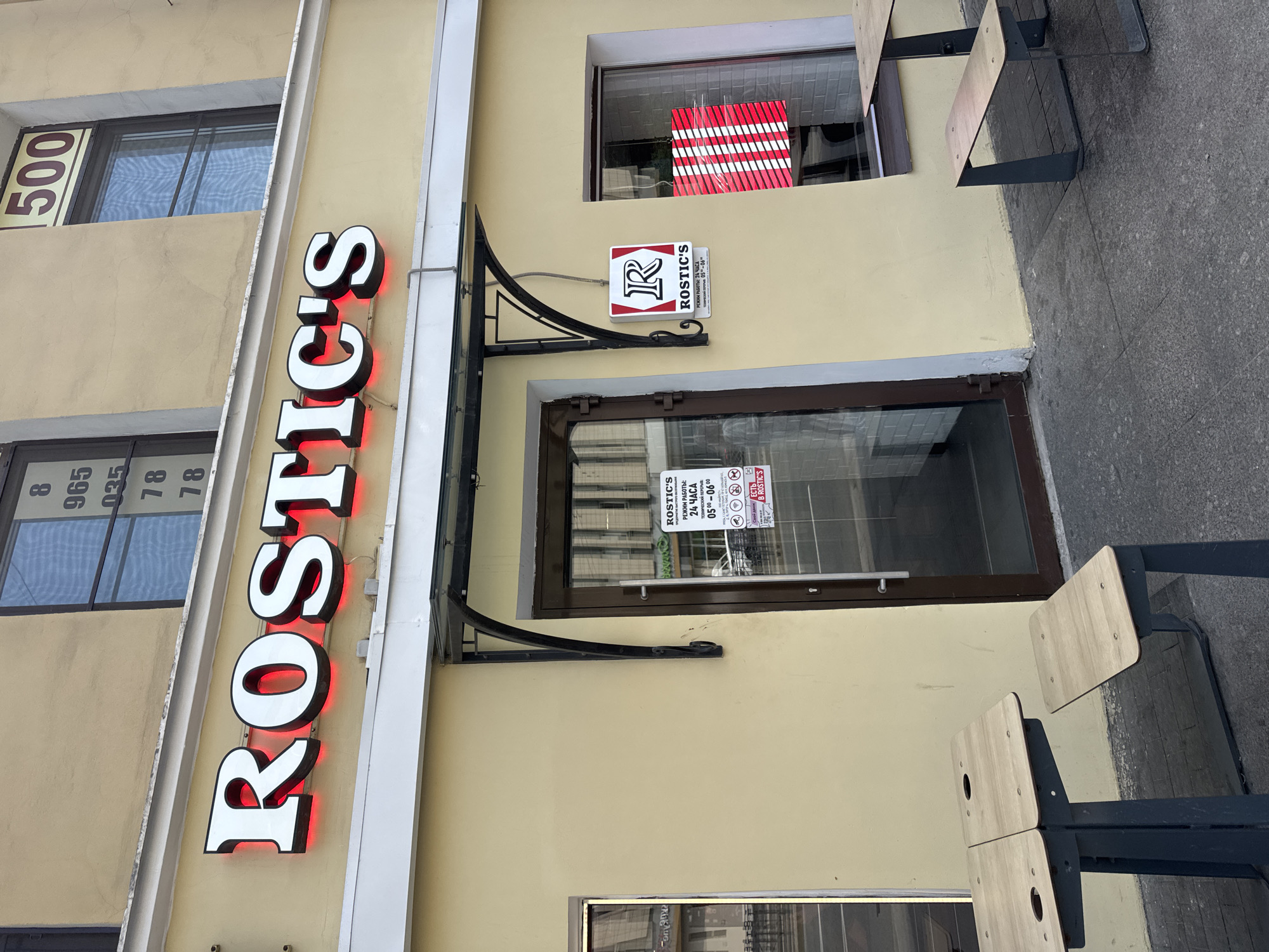 The image shows the facade of a business named "ROSTIC'S" in Saint Petersburg, Russia. The business name is displayed prominently in large, illuminated white letters with red backlighting, mounted above the entrance. The building's facade is painted a pale yellow.
To the left of the entrance, there is a window with a reflective surface. To the right of the entrance is a glass door, and to the right of the door, there is a window displaying a red and white striped pattern. Above the entrance, a dark metal awning extends outwards.
A sign with the "R" logo for ROSTIC'S is mounted on the wall next to the entrance. Beneath this logo, smaller text indicates business hours: "РЕЖИМ РАБОТЫ: 24 ЧАСА" (Work Schedule: 24 Hours) and "ТЕХНИЧЕСКИЙ ПЕРЕРЫВ: 05:00-06:00" (Technical Break: 05:00-06:00). There are also smaller signs with icons that appear to be related to rules or services.
Outside the building, in front of the entrance and to the left, are two wooden tabletops attached to dark metal legs, resembling outdoor seating. The ground in front of the establishment is paved with dark grey tiles. The overall scene depicts the exterior of a commercial establishment, likely a fast-food restaurant, given the name "ROSTIC'S" which is a known fast-food chain. The lighting suggests it is daytime.