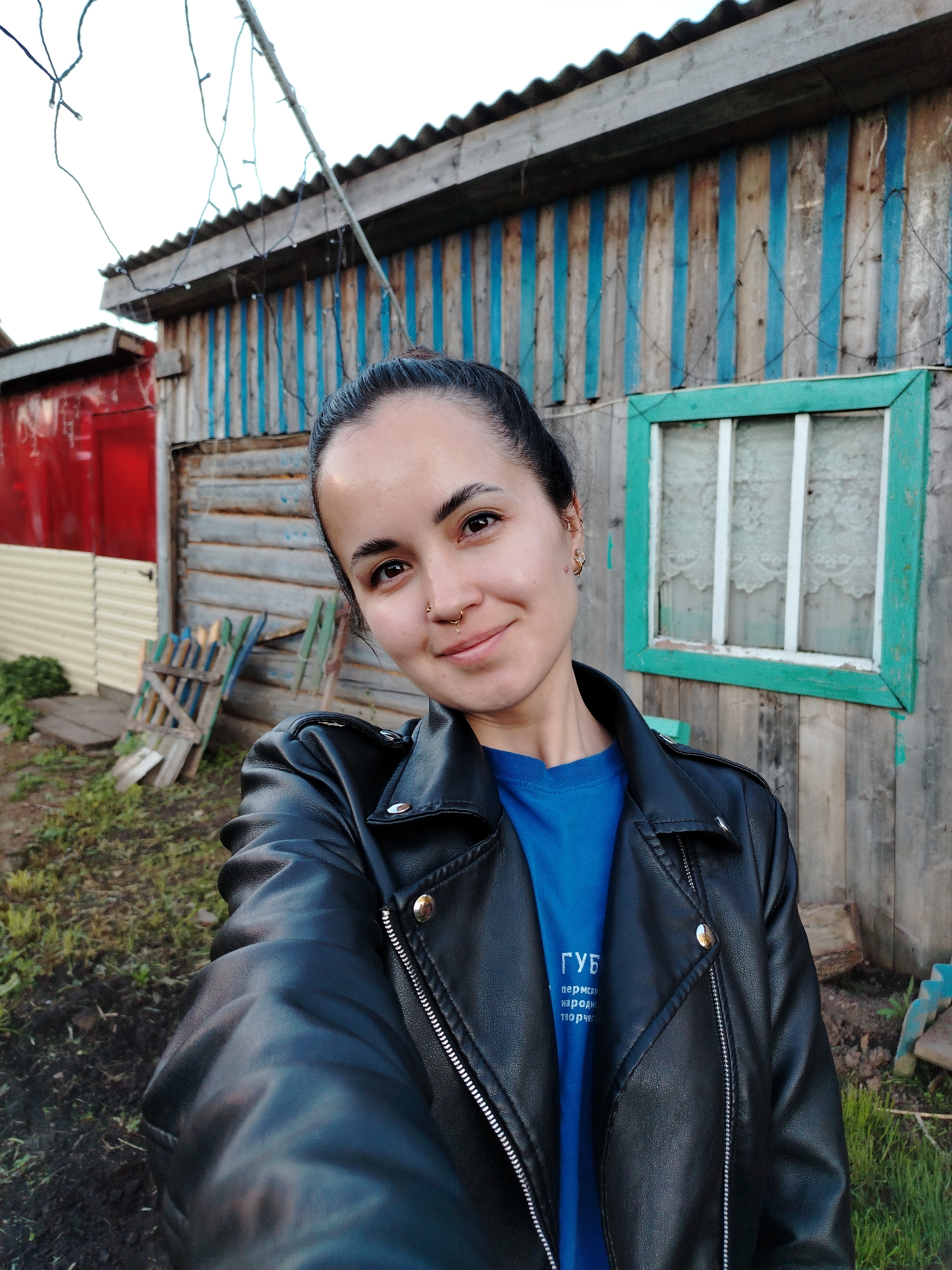 A woman with dark hair pulled back into a bun is the central subject of this outdoor selfie. She is wearing a black leather jacket over a blue t-shirt. Her facial features include a nose ring and earrings.
The background is a rustic, possibly rural, setting. There's a wooden structure with a blue and unpainted wood slat pattern, alongside a small window with a green frame. Adjacent to this is a red building with corrugated metal walls. Discarded wooden pallets are on the ground. The setting appears to be in the city of Гожан, Russia.