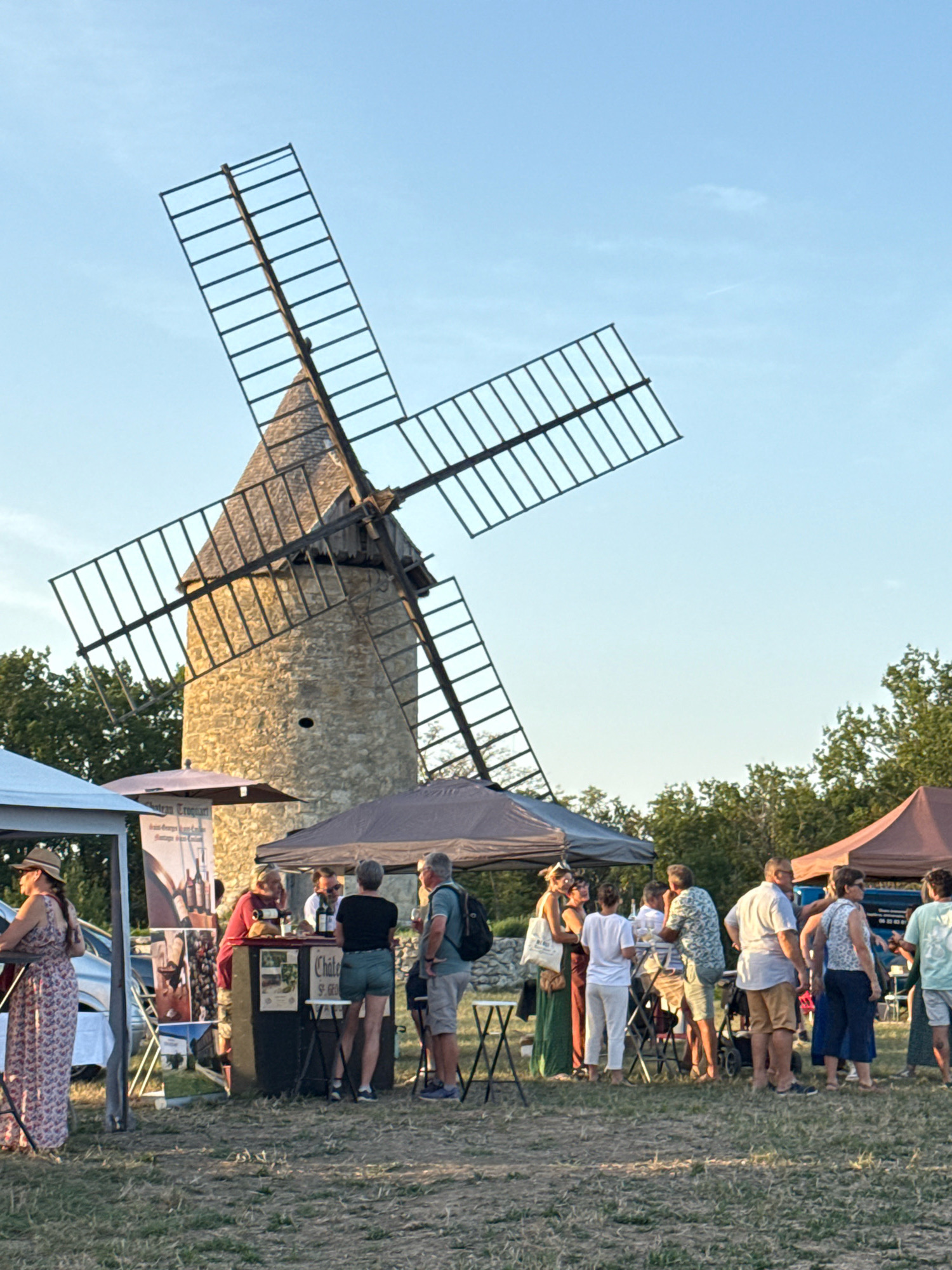 The image depicts an outdoor event taking place in front of a historic stone windmill in Montagne, France. The windmill, a prominent structure, has four large wooden sails against a clear blue sky. In the foreground, several tents and stalls are set up, suggesting a market or festival. A group of people are gathered around these stalls, some interacting with vendors, others socializing. They are dressed in casual summer attire. The ground is covered in dry grass, indicating a rural or park-like setting. Trees are visible in the background, surrounding the windmill. The lighting suggests it is late afternoon or early evening.