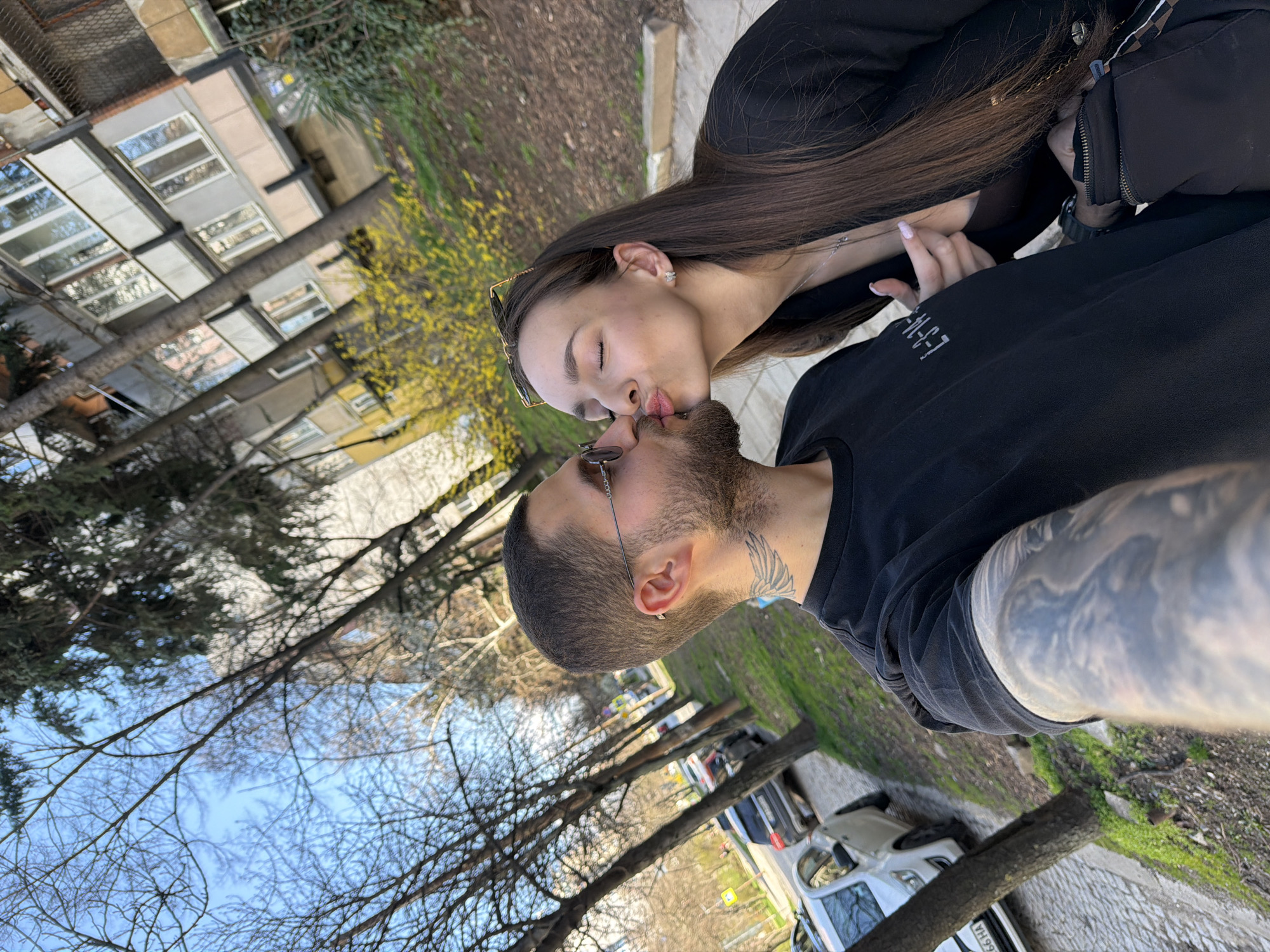 A man and woman are kissing in an outdoor setting. The man has a beard and a tattoo on his neck, while the woman has long, dark hair and is wearing glasses on top of her head. They are both dressed in dark clothing. The background shows a city street with trees, a building with windows, and parked cars. Based on visual clues such as the car license plate, the image was likely taken in Sofia, Bulgaria.