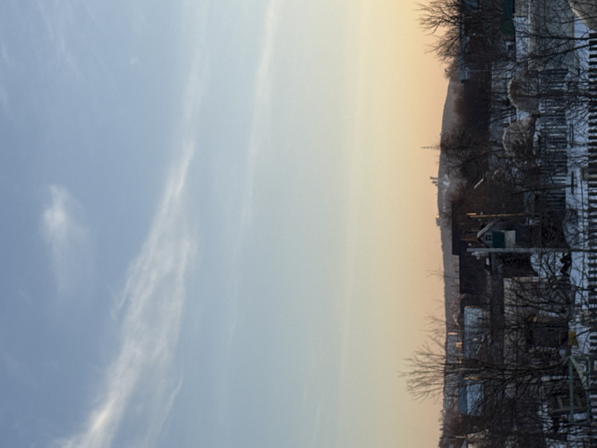 The image shows a view of Chaikovsky, Russia, during what appears to be a winter or early spring morning. The sky transitions from a light blue to a warmer, yellowish hue near the horizon, likely indicating sunrise. Thin, wispy clouds stretch across the blue portion of the sky. The foreground depicts a residential area with houses and bare trees. Snow is visible on the ground and rooftops. The scene is viewed from a slightly elevated position, offering a panorama of the cityscape.