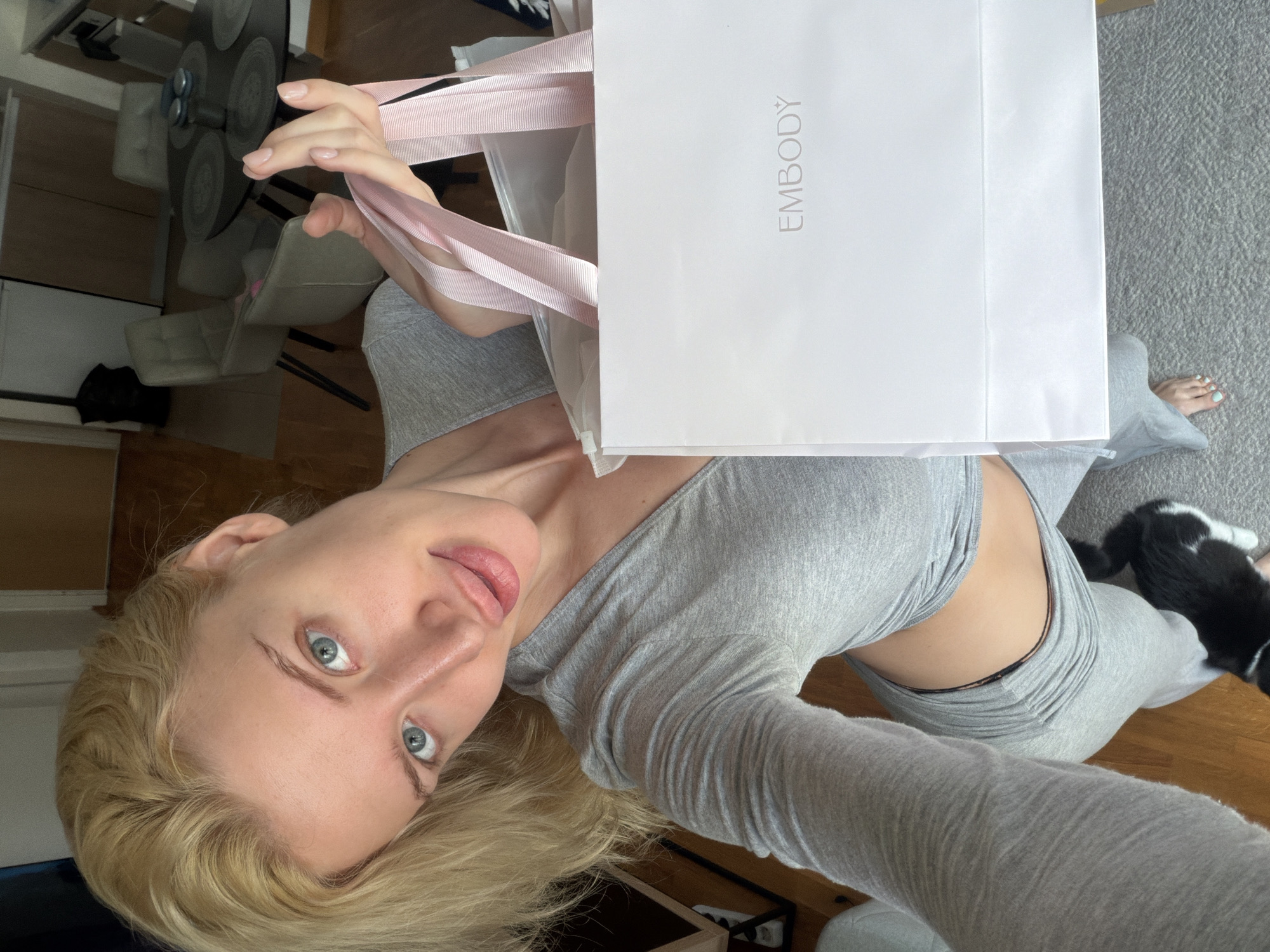 A woman with blonde hair and light eyes, wearing a light gray long-sleeved top and matching pants, holds a white shopping bag with the word "EMBODY" printed on it. The bag has pink ribbon handles. The woman appears to be in an indoor environment, possibly an apartment. A cat is partially visible in the lower right of the frame. The room contains furniture like a glass table and chairs. The location is possibly in Belgrade, Serbia.