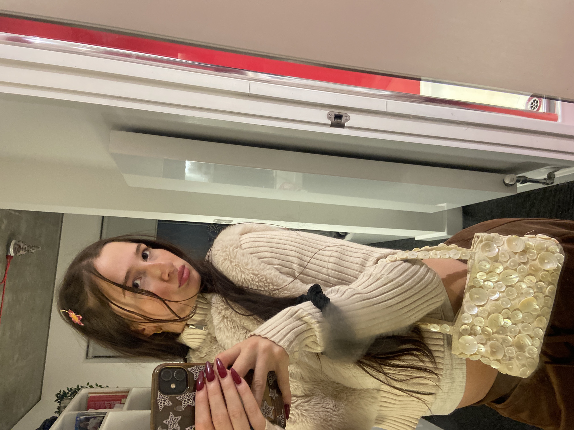 The image shows a young woman taking a selfie in a mirror. She is wearing a cream-colored sweater and holding a cellphone with a star-patterned case. She has long brown hair and is wearing a small flower clip in her hair. She is also carrying a small handbag covered in buttons.
The background is blurred, but it appears to be a room with white walls and shelving. There is a red stripe along the top of the mirror. The location is likely indoors and is not identifiable as being in London.