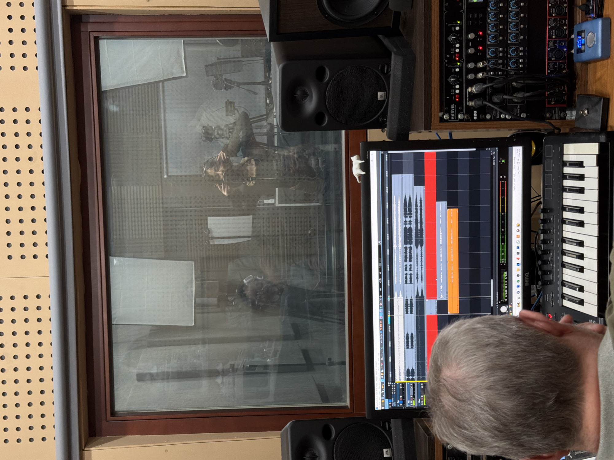 The image depicts a recording studio control room in Krasnoyarsk, Russia, with a clear view into an adjacent recording booth through a large glass window. In the foreground, the back of a person's head with gray hair is visible, looking intently at a computer monitor. The monitor displays a Digital Audio Workstation (DAW) interface, showing multiple colored audio tracks with detailed waveforms, indicating an active music production or editing session. A small white cat figurine is perched on the upper right side of the monitor, which has a "BenQ" logo.
To the right of the monitor, a black M-Audio MIDI keyboard controller is visible. Above the monitor, a rack of audio equipment is stacked, featuring various devices including a unit labeled "MARVEL" and another marked "828", along with a red Focusrite Scarlett audio interface. Numerous cables connect these devices. A JBL studio monitor speaker is positioned to the left of the equipment rack. The wall to the left of the window is covered with beige perforated acoustic paneling.
Through the window, a reflection of the control room interior is visible, but also partially discernible is a person inside the recording booth. This individual appears to be wearing headphones and is positioned near a microphone on a stand, suggesting they are in the process of recording vocals or an instrument. The booth's interior walls show acoustic treatment, with some areas covered by white sheets of paper. The scene captures a professional audio production environment.