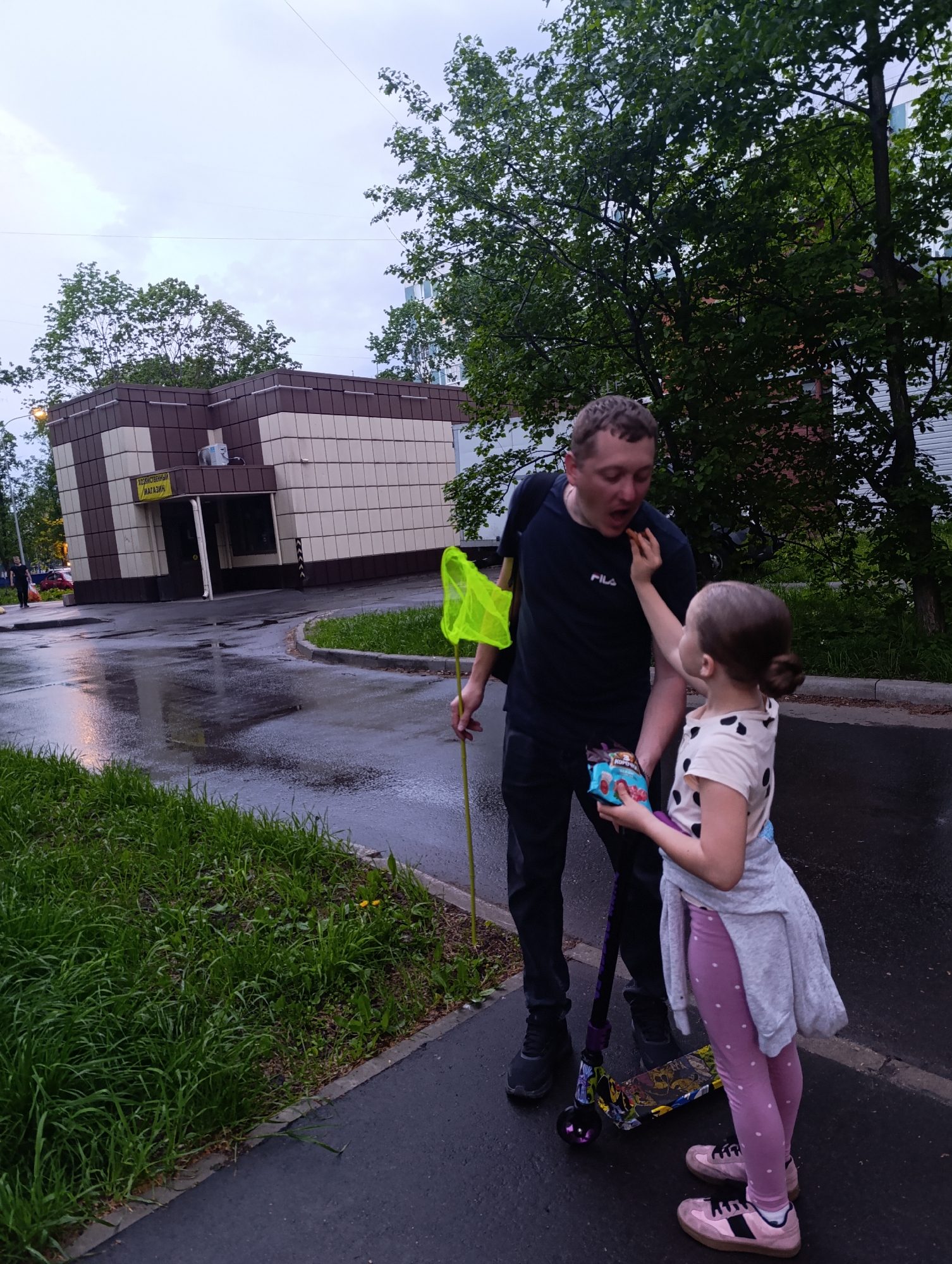 The image depicts a man and a young girl interacting on a wet city street. The man, wearing a dark blue shirt and dark pants, is holding a yellow net and has a colorful scooter leaning against his leg. The girl, dressed in a polka dot shirt, pink leggings, and a gray jacket, is feeding him a snack from a small bag she's holding. They are standing on a sidewalk next to a grassy area. Behind them, there is a building with beige and brown panels. The street appears wet, suggesting it has recently rained. The background includes trees and another building further away. The location is Unknown, Unknown.