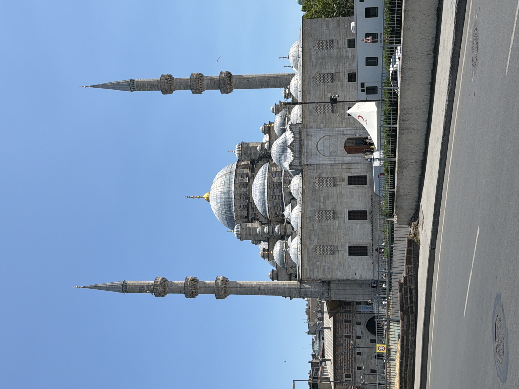 The image shows the Yeni Cami (New Mosque) in the Eminönü district of Fatih, Istanbul, Turkey. The grand mosque features a central dome, flanked by smaller domes and supported by stone minarets. The building is constructed from light-colored stone, and its architectural details are intricate. In front of the mosque, there is a street with a sidewalk and a low concrete wall. Beyond the wall, a metal fence encloses an area with some people, a white tent with a red crescent logo, and a white mobile unit. Trees are visible on the right side of the image, and the sky is clear and blue.