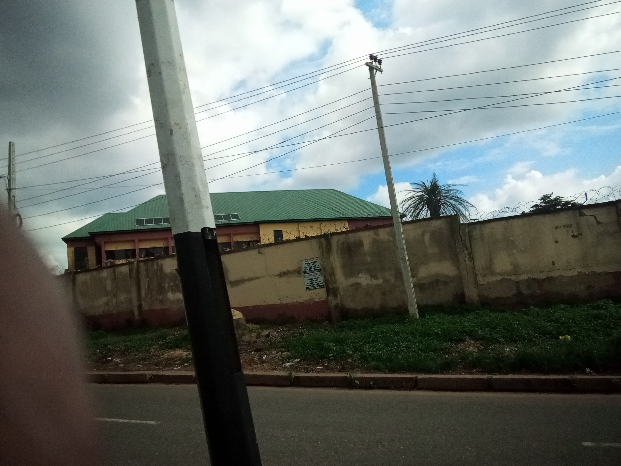 The image shows a view of a building with a green roof and yellow walls, enclosed by a concrete wall topped with barbed wire. The building appears to be located in Nibo, Nigeria, based on the context provided. In the foreground, a portion of a large, white, octagonal pole is visible, obscuring a part of the scene. Electrical wires are strung across the frame, supported by utility poles. The ground in front of the wall is covered with green grass and some debris. The sky is cloudy with patches of blue. The scene is captured from a street level perspective, with a portion of an asphalt road visible at the bottom of the image.