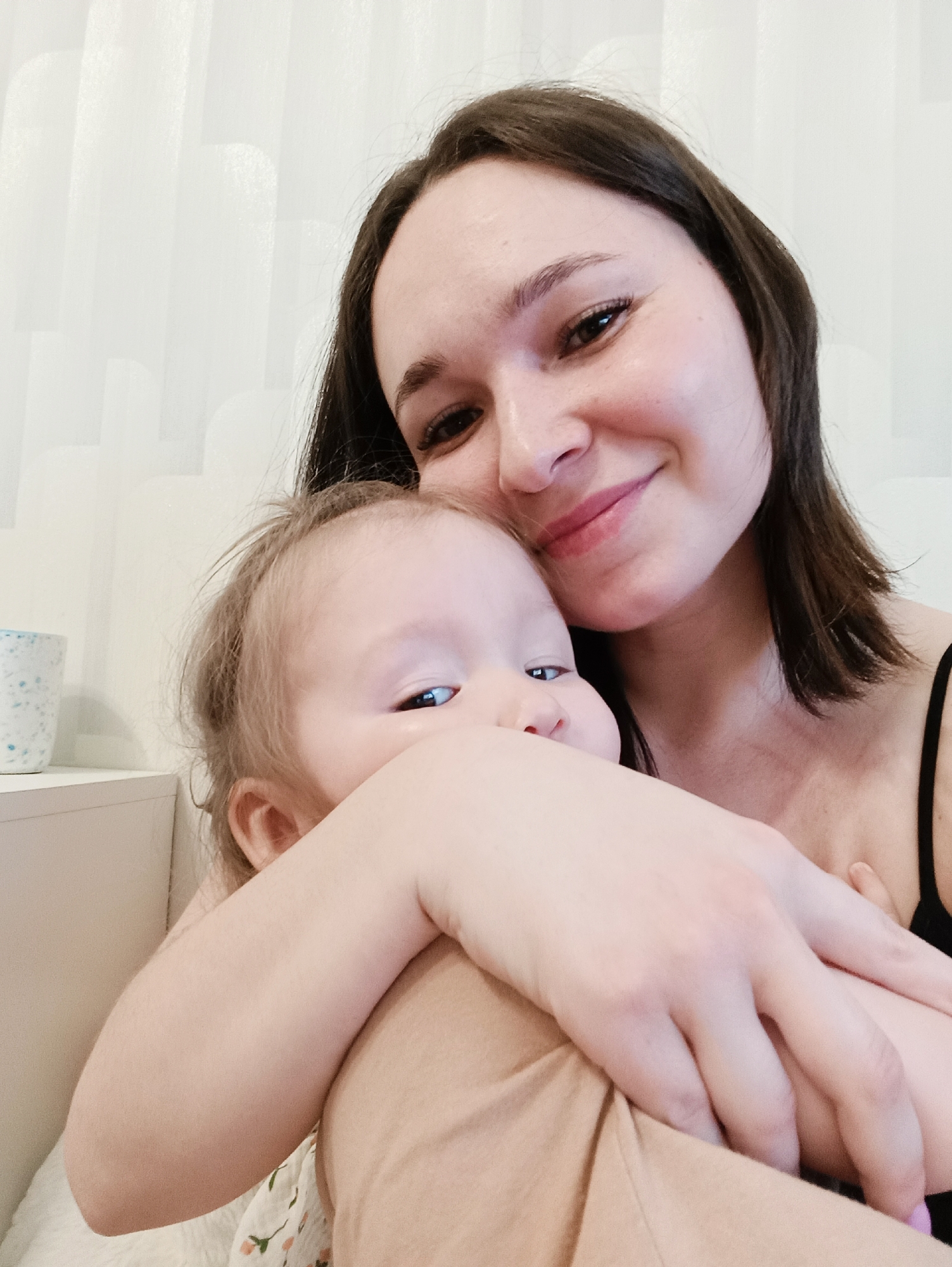 The image displays an adult female embracing an infant in an indoor setting. The adult, positioned on the right, has dark brown hair and light skin, with her visible right eye and mouth indicating a smiling expression. She is wearing a black spaghetti strap top. Her left arm is wrapped around the infant. The infant, on the left, has light hair and light skin, with only the left side of its face, including the left eye, clearly visible. The infant appears to be wearing light-colored, possibly beige or tan, clothing with a subtle floral pattern. In the upper left background, a white or light-colored speckled mug is placed on a white surface, likely a shelf or nightstand. The background consists of a plain white wall or vertical blinds. The specific location within Izhevsk, Russia, cannot be determined from the visual information provided.
