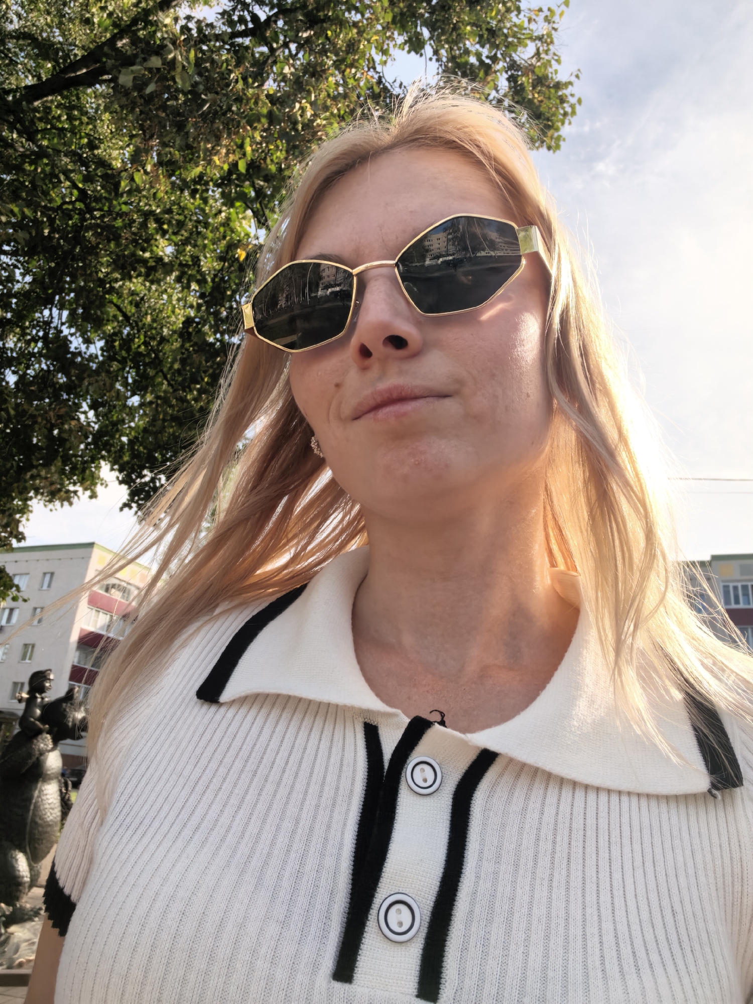 A young woman with blonde hair and sunglasses is standing outdoors, possibly in Sterlitamak, Russia, based on the presence of Soviet-era apartment blocks in the background. She is wearing a white ribbed polo shirt with black trim and buttons. In the background, a tree with green leaves and a statue of a mythical creature are visible. The sky is clear and bright, with the sun casting a warm glow. The woman is looking at the camera, and her expression is relaxed.