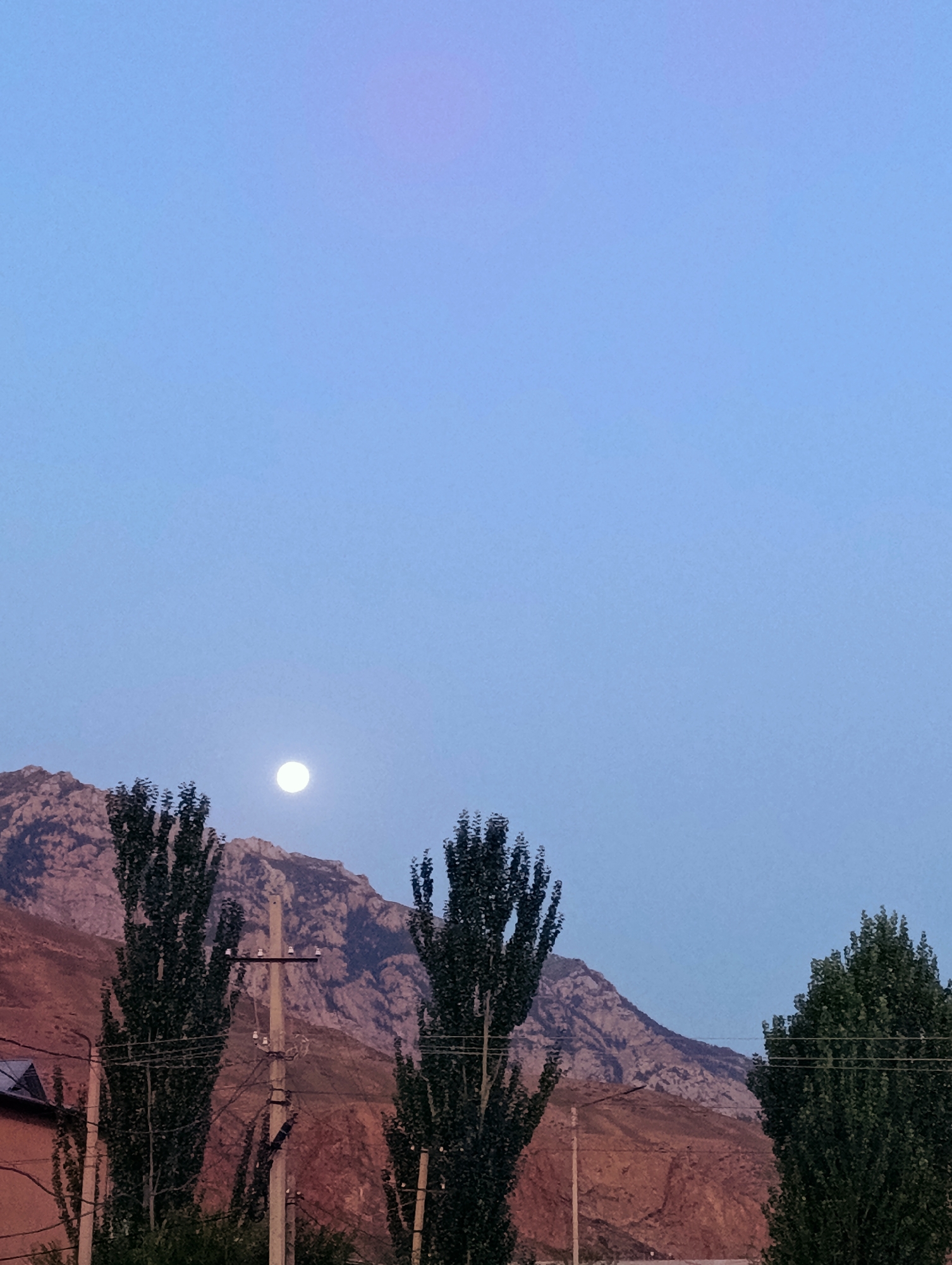 The image captures a twilight scene in Naryn City, Kyrgyzstan, with a clear, pale blue sky dominating the upper portion. A full moon is visible in the upper left quadrant of the sky, emitting a soft glow. Below the sky, a mountain range stretches across the background, its rocky slopes appearing in shades of brown and gray. In the foreground, three tall, slender trees with dark green foliage stand prominently. A utility pole with visible wires and insulators is positioned between the two left trees. To the far left, a portion of a building with a dark roof is visible. The overall impression is a serene, natural landscape at dusk.