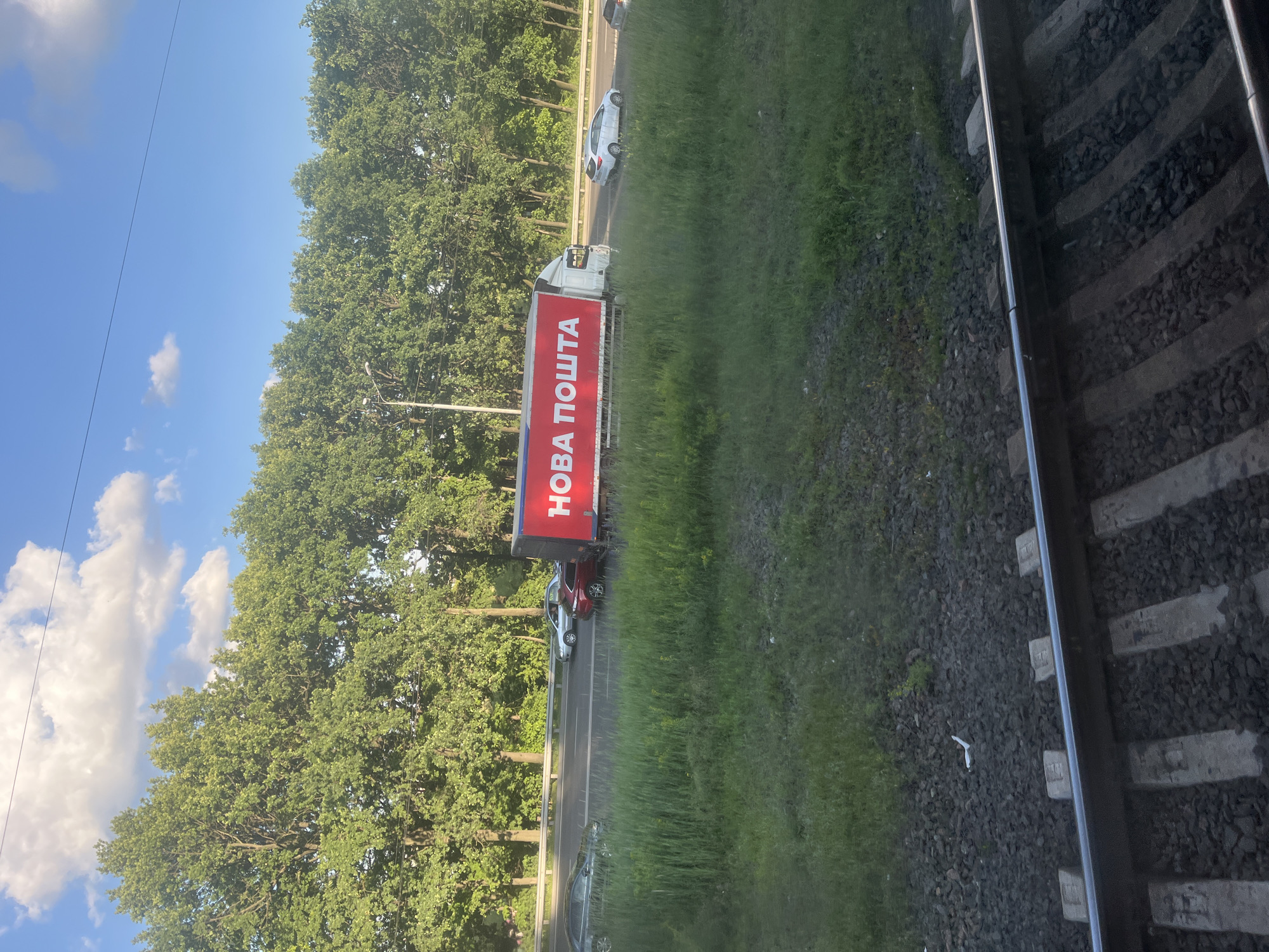 The image shows a street scene on a sunny day in Kyiv, Ukraine. In the foreground, there are train tracks. Beyond the tracks, there is a grassy area with tall grass. In the middle ground, there is a highway with traffic. A large red and white truck with the text "НОВА ПОШТА" (Nova Poshta) is visible on the side of the trailer, traveling on the highway. Several cars are also on the highway. In the background, there is a dense line of green trees. The sky is blue with some white clouds.