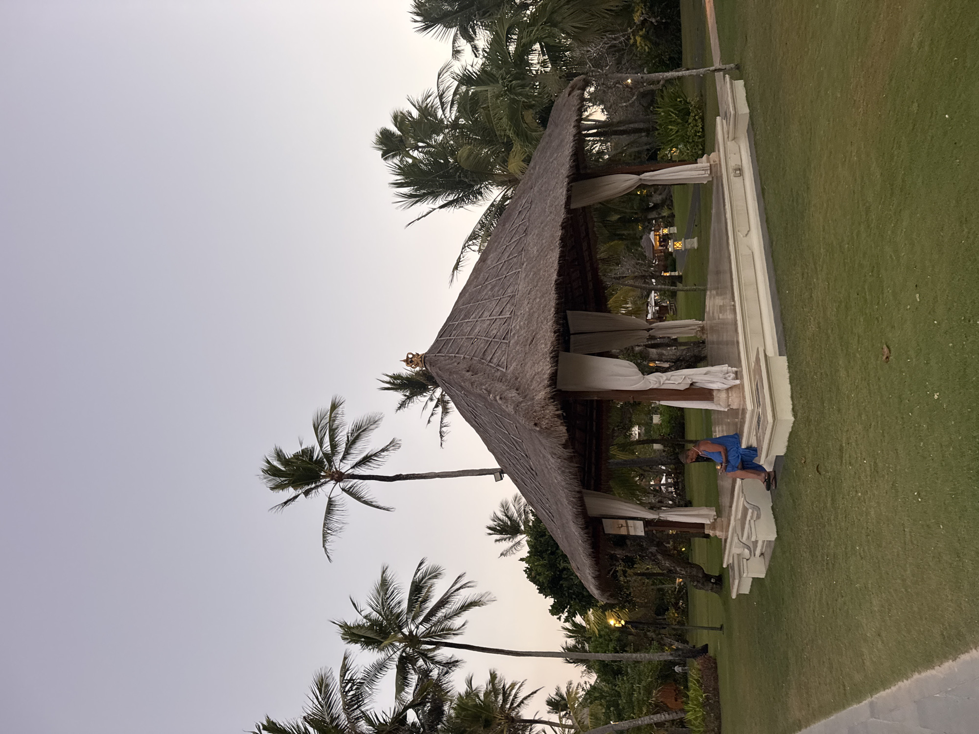 A woman in a blue dress sits on the raised platform of a thatched-roof pavilion in Kuta Selatan, Indonesia, facing left and looking down at her phone. The pavilion has white drapes hanging from its sides. The pavilion is situated on a well-maintained green lawn, with several palm trees visible in the background. The sky is a pale, twilight hue. A paved path runs along the left edge of the frame, and another path can be seen winding through the lawn in the distance. Several outdoor lanterns are also visible in the background.