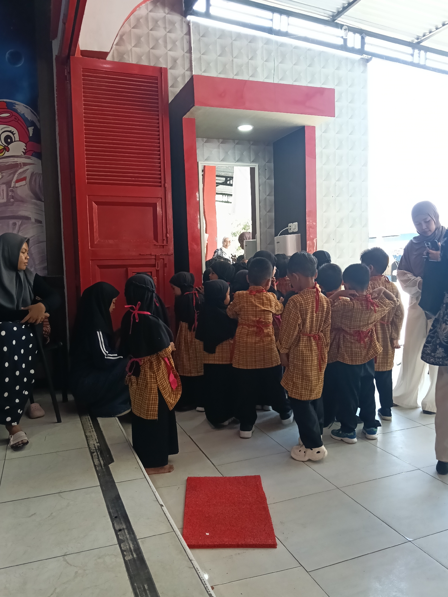 The image depicts a group of young children, likely students, gathered in an outdoor or semi-outdoor area. The children are dressed in matching yellow and orange plaid outfits with red sashes tied around their waists, suggesting a uniform or costume for an event. They are facing away from the camera, with their backs towards an entrance or doorway framed by red and white structures. The environment appears to be an educational institution or a public space with a tiled floor. Several adults, presumably teachers or guardians, are present, observing or interacting with the children. One adult woman in a hijab is seen to the left, seated and looking towards the group. Another adult woman in a hijab is visible in the background, partially obscured. The area features a red double door and a mural on the left wall depicting a cartoon character in an astronaut suit. The overall impression is of an organized gathering of children, possibly for a school activity or celebration, in a location in Indonesia.