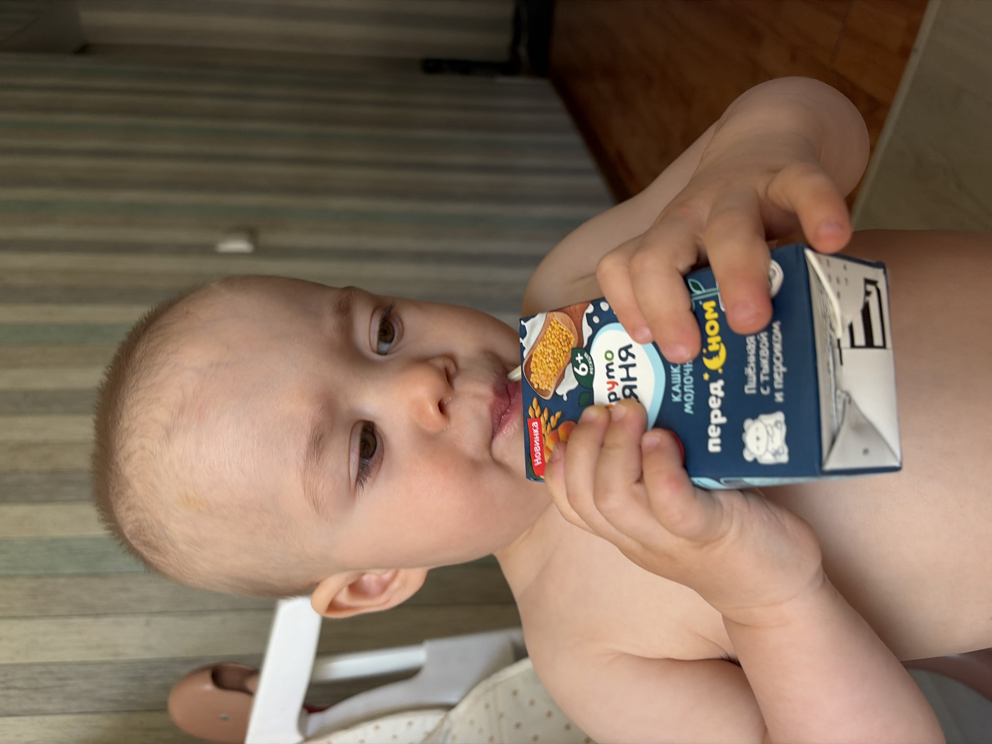 A baby is drinking from a juice box. The juice box is labeled in Russian. The brand of the juice box is "Fruto Nyanya". The baby is shirtless. The baby has blonde hair. The background is blurry, but appears to be a room with wooden floors. The image was taken in the city of Chaikovsky, Russia.