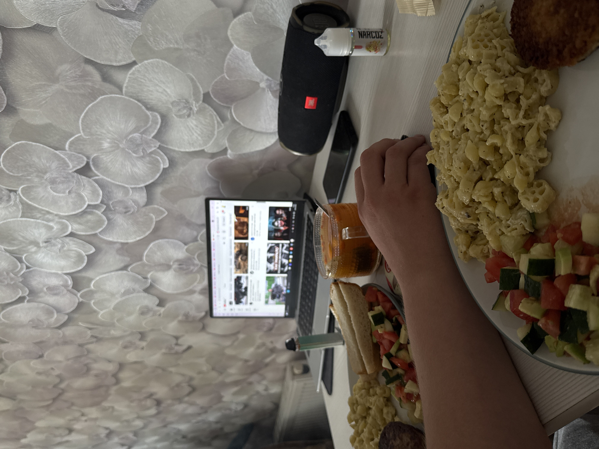 The image shows a table setting with various items. A person's arm is visible near a plate of food consisting of pasta, a patty, and a tomato-cucumber salad. Next to the plate, there is a sandwich and a glass of orange liquid. A laptop displays a webpage with thumbnails of videos. To the left, a black JBL speaker and a bottle labeled "NARCOZ" are placed. The table surface is white. The wallpaper features a white floral pattern. The location in the city of Shirkovo, Russia, cannot be confirmed from the provided image.