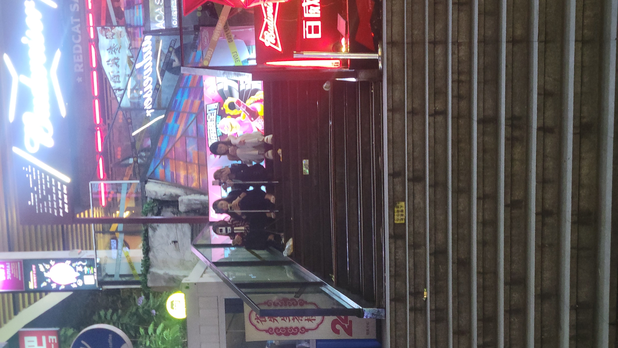 Here is a detailed description of the image:
**General Scene:**
The image depicts an outdoor scene in an urban area, likely at night, given the presence of neon signs and the lighting. The setting appears to be a street or pedestrian area with stairs leading up to an entrance. There are a lot of billboards with neon lights.
**People:**
There are a few people visible on the stairs. They appear to be a group of young girls, and they may be posing for pictures, as at least one of them has a phone in their hand.
**Objects:**
* **Stairs:** A set of concrete stairs occupies a significant portion of the image.
* **Neon Signs/Billboards:** Numerous brightly lit neon signs and billboards are visible in the background. One sign features a Budweiser logo.
* **Glass Partition:** A glass partition or railing separates the stairs from the adjacent area.
* **Signage:** A banner or sign with Chinese characters is attached to the glass partition near the bottom of the stairs.
**Environment and Action:**
The atmosphere is vibrant and energetic, typical of a bustling urban nightlife area. The people on the stairs are likely taking photos in front of the colorful billboards and neon signs.
**Location:**
Although it is not possible to determine the location without further information, it is likely an East Asian city, given the signage and the style of the architecture.