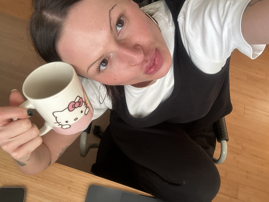 A selfie, taken from above, features a woman holding a Hello Kitty mug. She has dark hair and fair skin, wearing a white t-shirt layered with a black top. Large hoop earrings are visible. She is seated, wearing black pants on what looks like an office chair. A wooden desk is visible in the foreground, with a smartphone and laptop placed on it. Her nails are painted black. The image is brightly lit.