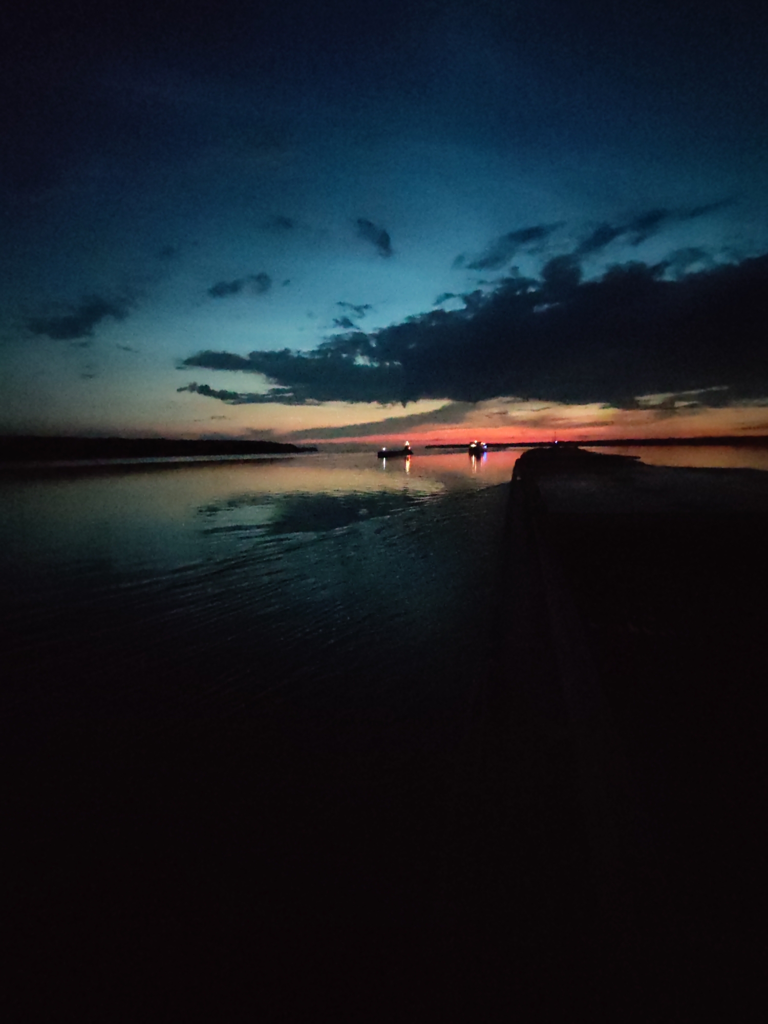 The image captures a serene twilight scene on the water, possibly the Gulf of Finland, near Kamenniki, Russia. The sky transitions from a deep, dusky blue overhead to hues of orange and pink near the horizon, indicating the last moments of sunset. Dark, scattered clouds drift across the upper sky.
The water in the foreground is dark and reflective, showing subtle ripples. In the mid-ground, two small boats with lights illuminated are visible on the water, suggesting they are either fishing or transiting the area. A long, dark, man-made structure, possibly a pier or breakwater, extends into the water from the right side of the frame, creating a stark contrast with the lighter horizon. The far shore is a dark, indistinct silhouette against the colored sky. The overall impression is one of quietness and solitude as night descends.