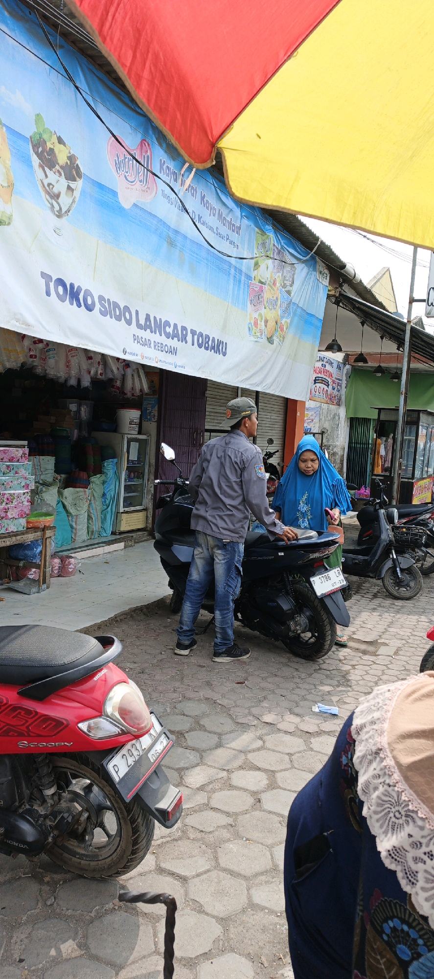 The image depicts an outdoor scene in front of a shop with a banner that reads "TOKO SIDO LANCAR TOBAKU PASAR REBOAN". A man wearing a gray shirt and jeans is standing next to a black motorcycle, seemingly interacting with a woman in a blue hijab. The woman is holding a piece of paper. The shop appears to sell various goods, with shelves stocked with items visible behind them. In the foreground, the rear of a red scooter is visible. The ground is paved with hexagonal stones. The environment suggests a marketplace setting in Kasiyan, Indonesia, with a red and yellow awning providing shade.