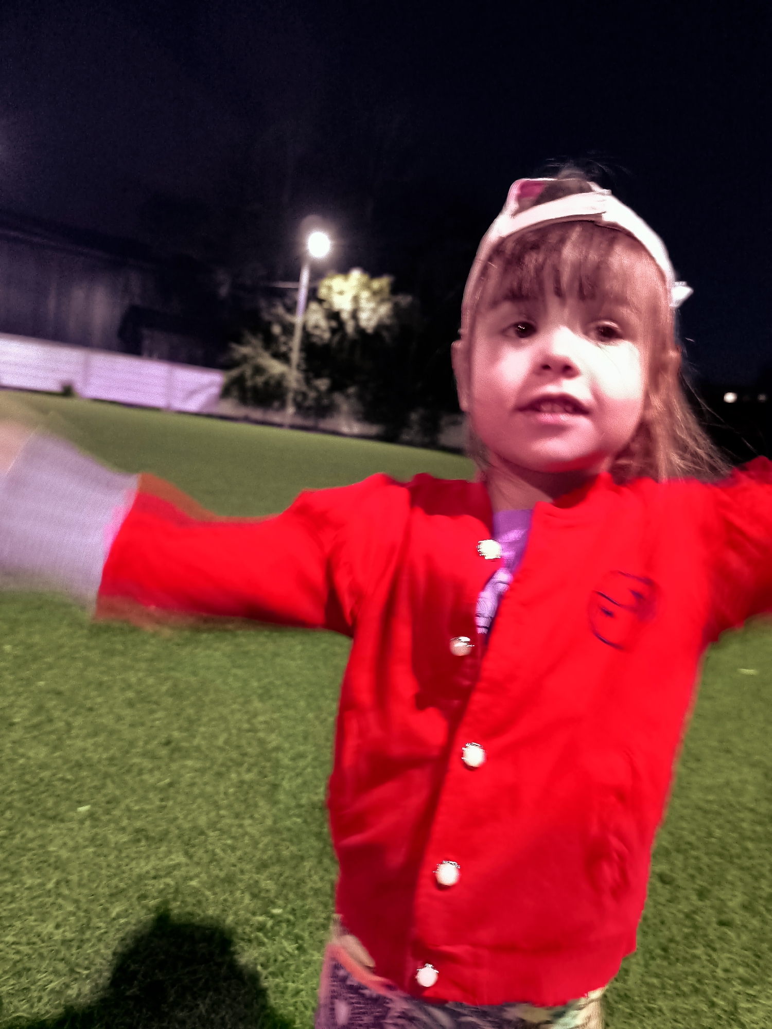 The media file depicts a young child, likely a girl, in a close-up, blurry shot taken at night. She is wearing a bright red cardigan over a purple shirt and patterned leggings. The child is facing the camera with her arms outstretched, suggesting she is in motion or playing. A white headband with pink accents is visible on her head. The background shows a green, grassy area, possibly a field or park, illuminated by a distant street light. In the far background, a building or fence can be seen. The lighting indicates it is nighttime, and the overall atmosphere is somewhat playful, given the child's pose. There are no discernible text elements in the image.