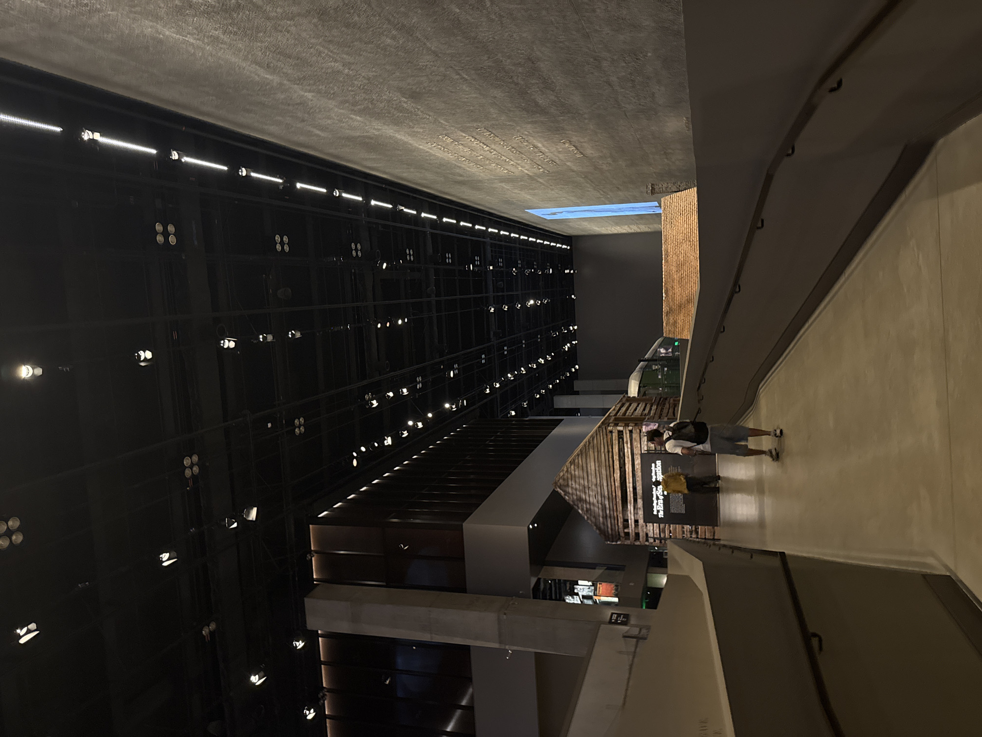 The image shows the interior of a museum in Washington, D.C. In the foreground, a ramp curves to the right, with a dark gray wall on the right side and a lighter gray wall on the left, both featuring dark handrails. Further into the museum, a rustic wooden structure resembling a cabin or small house is visible. Behind the wooden structure, a sign reads "Freedom's Footprints The Era of Segregation." To the left of the wooden structure, a dark-colored building with vertical windows is partially visible. In the distance, a large, dark-textured wall rises, with what appears to be text or an inscription on its surface. The ceiling is black and features a grid of lights, with some lights appearing as clusters of circular fixtures and others as linear LED strips. Two individuals are present in the scene: one person walking away from the camera on the ramp, wearing a backpack and shorts, and another person standing near the wooden structure, wearing a yellow shirt. The lighting is dim, creating a somber atmosphere, typical of a museum exhibit.