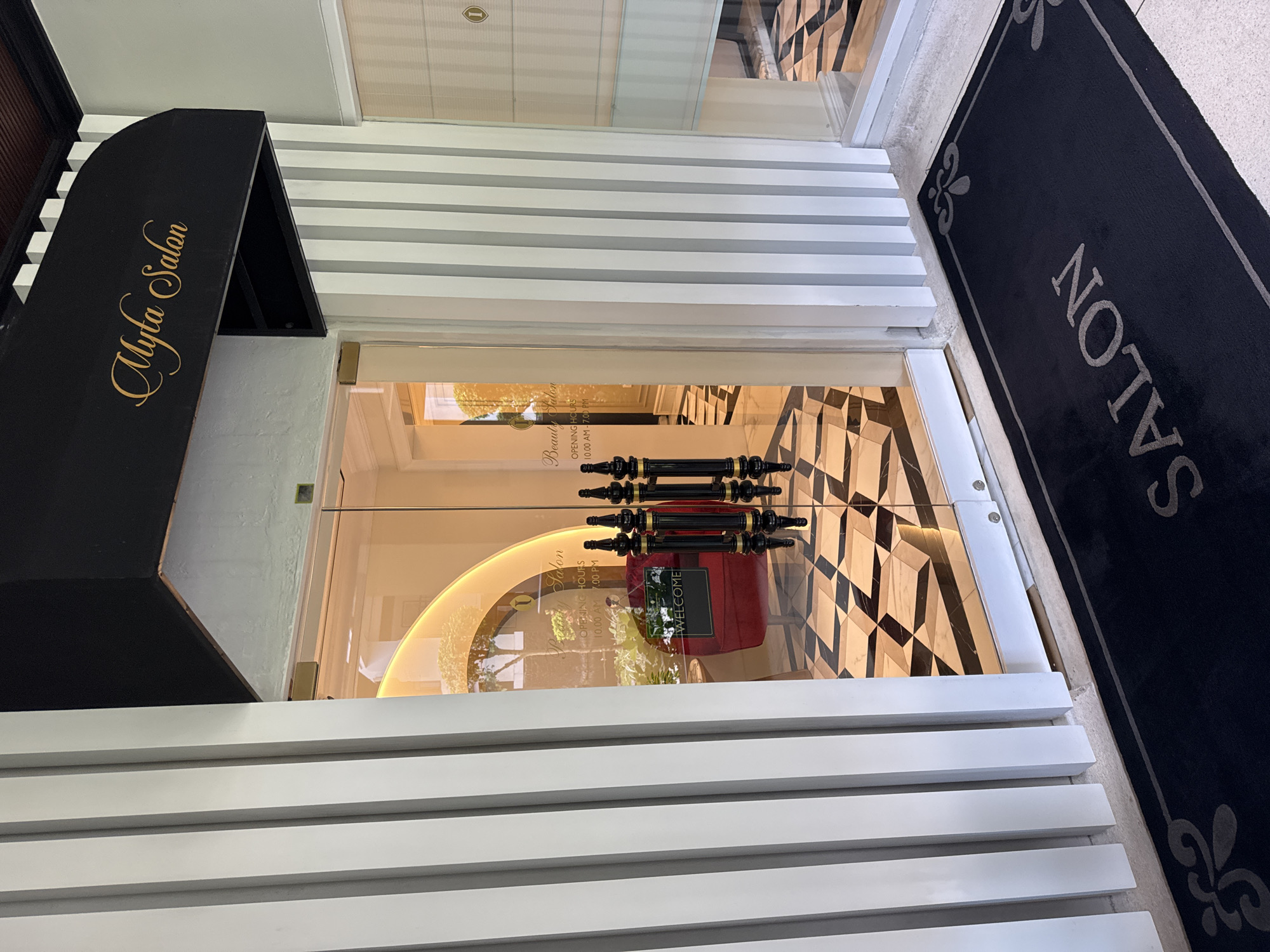 The image captures the entrance of "Myla Salon," situated in Kuta Selatan, Indonesia. The entrance features double glass doors with ornate black and gold handles. Inside, a red upholstered chair is visible, along with an arched decorative feature and a "Welcome" sign. A black doormat with "Salon" inscribed on it lies outside the entrance. The building facade has horizontal white and gray stripes. The awning above the door is black with the salon's name in gold script. The opening hours, "10.00 AM - 7.00 PM," are displayed on the glass door. The floor visible inside the salon has a black and gold geometric pattern.
