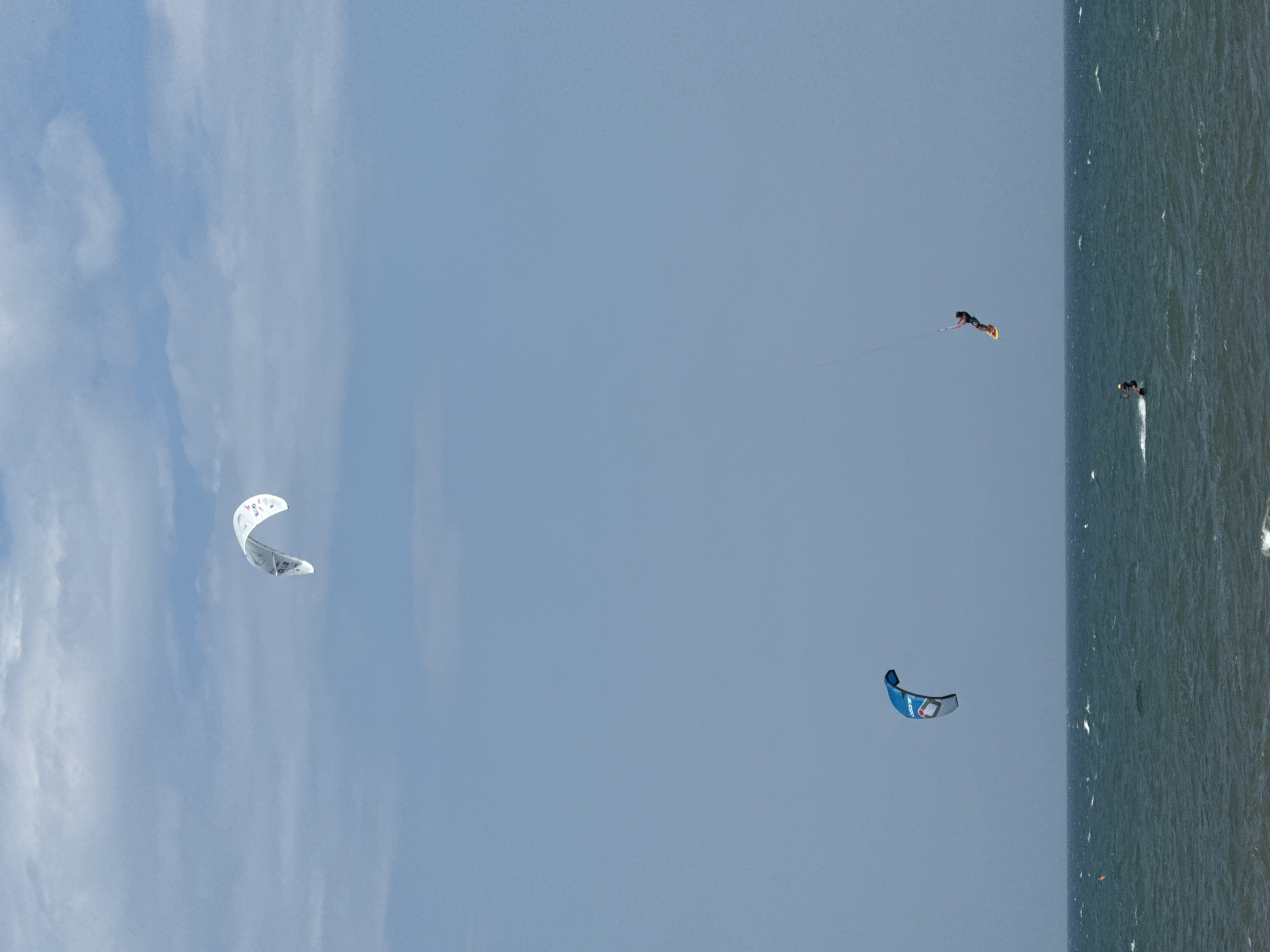 The image depicts a wide view of the ocean with a clear blue sky containing sparse clouds. Three kite surfers are visible. One kite surfer is in the upper portion of the frame, with a white kite. Another kite surfer, with a blue and gray kite, is in the middle-left of the frame. The third kite surfer is in the lower right portion, mid-air above the water, attached to a kite that is mostly out of frame. A fourth, smaller figure is visible in the water in the lower portion of the frame, suggesting another individual engaged in water sports. The scene is consistent with the popular kite surfing conditions found at Mui Ne beach in Phan Thiết, Vietnam, which is known for its strong winds and expansive coastline. The sea is moderately choppy with whitecaps.