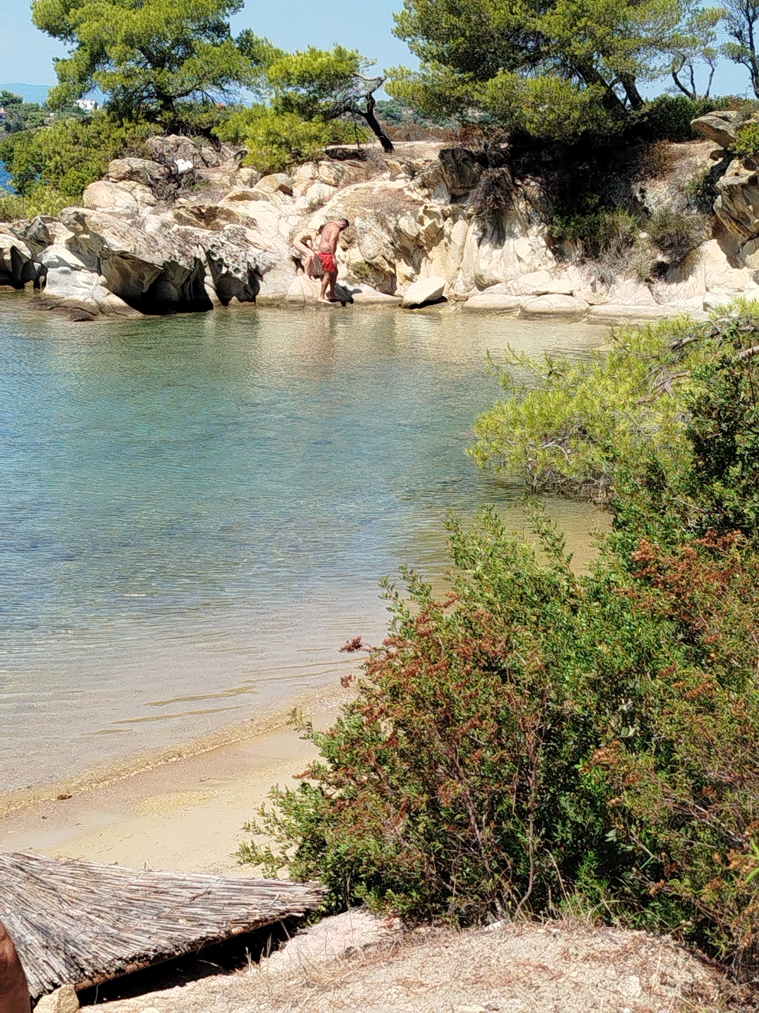 In Sithonia Municipal Unit, Greece, a man in red swim shorts is standing on rocky terrain near the water's edge, with his body bent over as if examining something on the rocks. He is on a small section of a rocky shoreline, with clear, shallow, turquoise water lapping at the base of the rocks. Behind him, the rocky landscape is covered with sparse green vegetation and trees. The beach area consists of sand and pebbles. In the foreground, a thatched mat or sunshade is visible, partially covering the sandy ground. The scene depicts a sunny day at a natural, coastal environment.