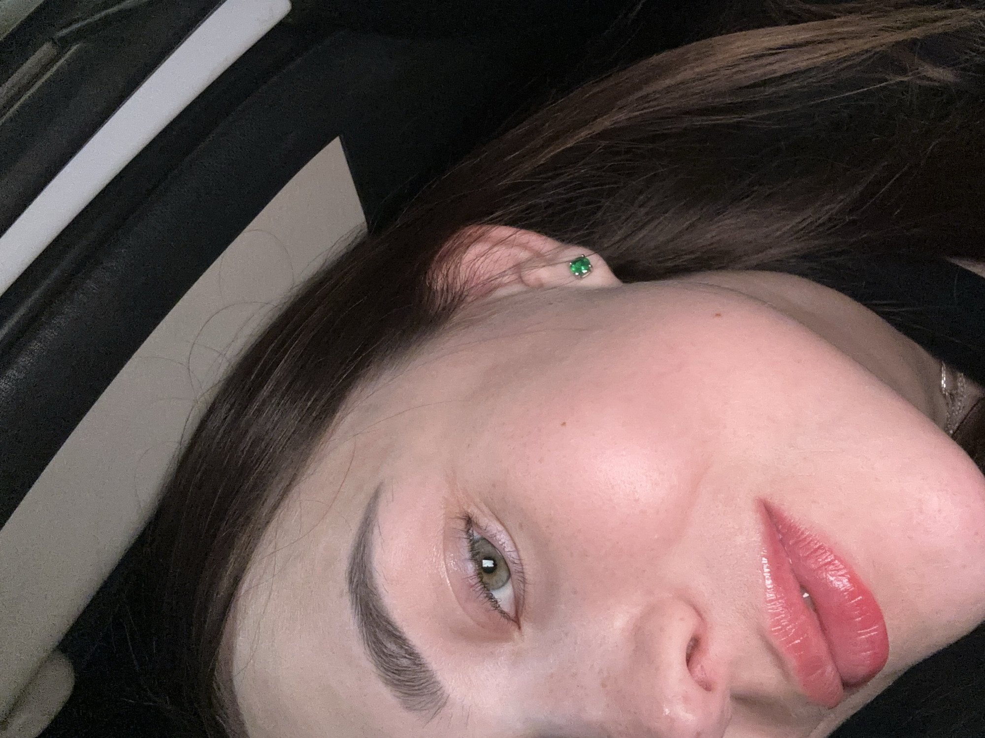 A close-up, low-angle, indoor shot shows the right side of a young woman's face. She has fair skin with a slight pink flush, prominent eyebrows, and green eyes. Her lips are glossy and a vibrant pinkish-red color. She is wearing a small, square emerald stud earring in her right ear. Her dark brown hair is visible, framing her face. The background is dark and out of focus, with a hint of a light-colored, curved object at the top. The image does not contain any elements that suggest a specific location within the city of Perm, Russia.