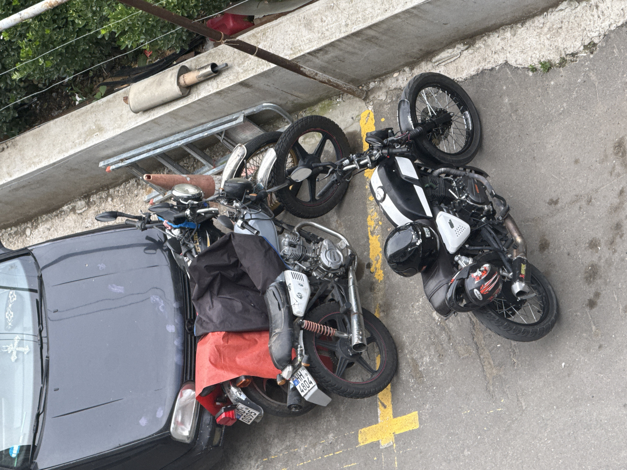 The image depicts an outdoor urban scene, likely in Tbilisi, Georgia, given the presence of Cyrillic script on a license plate and the architectural style. The foreground is dominated by two parked motorcycles. The motorcycle in the bottom right is black and white with racing stripes and has two helmets on it, one black and one red and black. The other motorcycle, parked behind it and slightly to the left, is silver and black with a red seat cover and a black tarp covering part of it. A portion of a dark-colored car is visible in the upper left corner. The motorcycles are parked on a paved surface marked with yellow lines, suggesting a parking area. To the right, a concrete wall runs along the scene, with a metal ladder leaning against it and a weathered exhaust pipe resting on top. Green foliage is visible beyond the wall. The overall impression is one of a parked vehicle and motorcycle area in an urban environment.