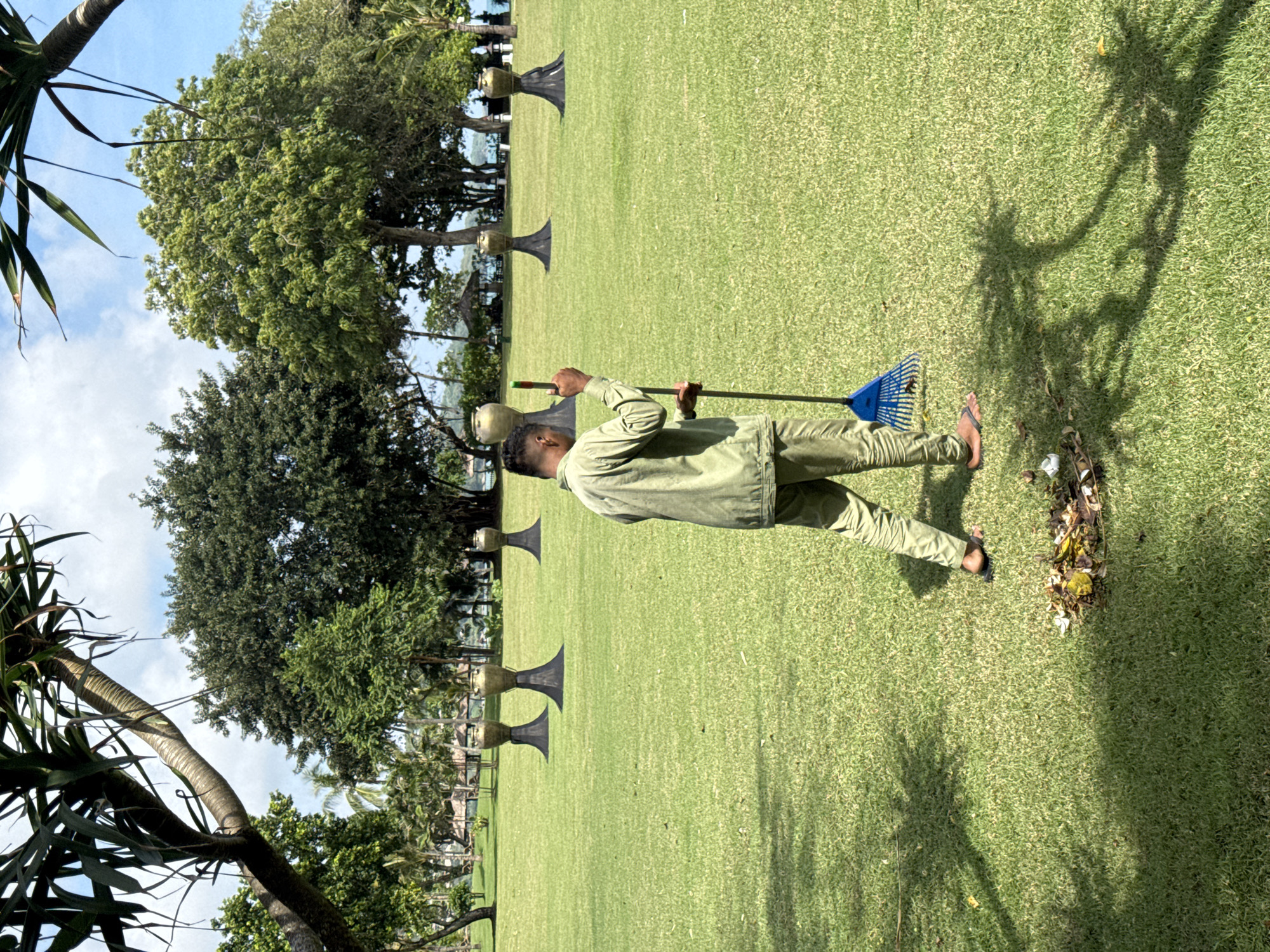 A man in light green clothing is raking leaves on a grassy lawn in Kuta Selatan, Indonesia. The location is characterized by several decorative urn-like structures spaced along the lawn's edge. Trees and palm fronds are present, casting shadows on the grass. The man is barefoot and using a blue rake. A pile of gathered leaves is visible near his feet. The sky is partly cloudy.