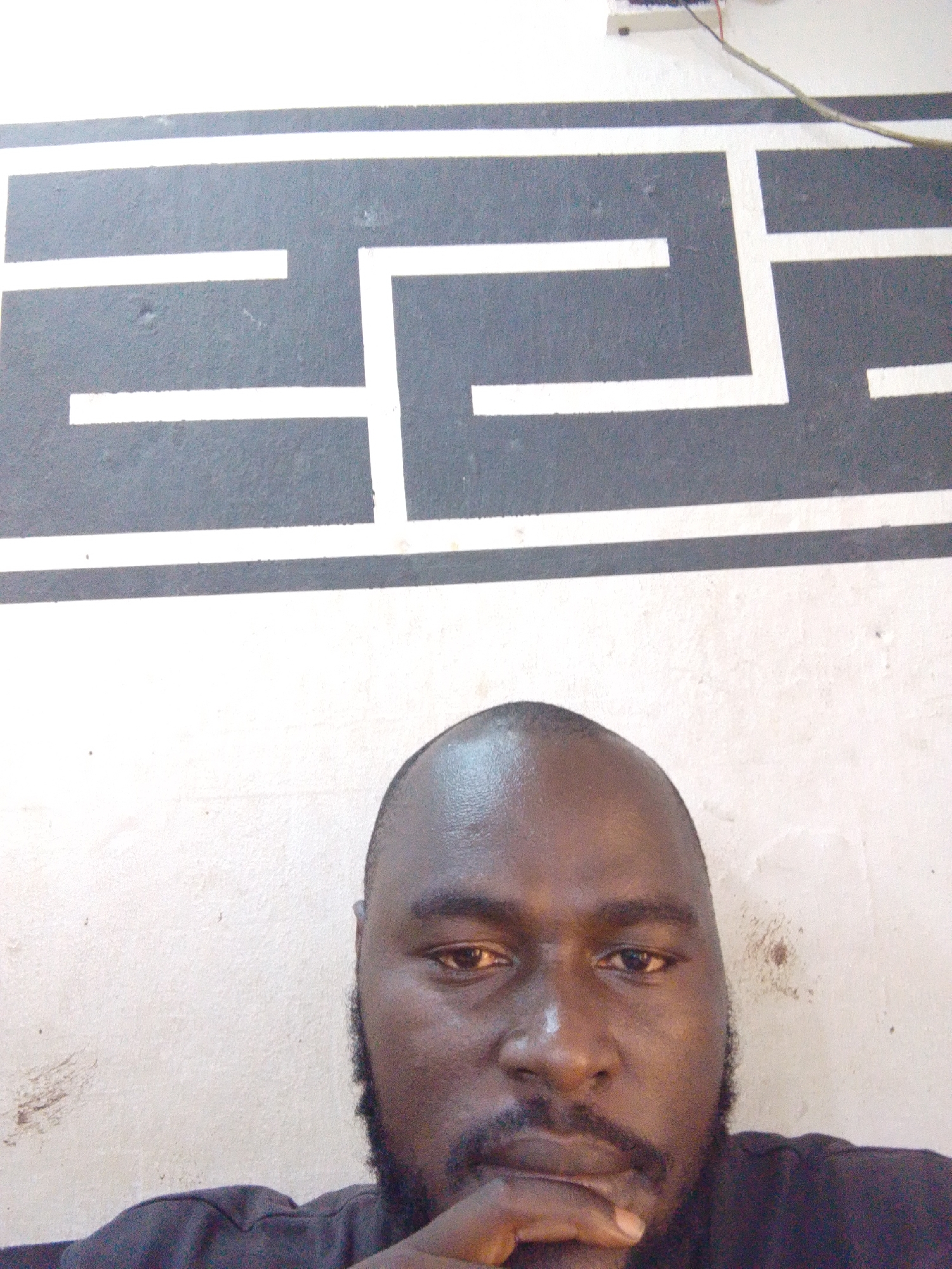 A close-up photograph depicts a Black male with a shaved head and a full beard, resting his chin on his hand. The background features a wall with a grey and white geometric design. A white electrical outlet box with wiring is visible in the upper right corner. The setting is likely indoors, given the wall and electrical fixture. There are no obvious indicators to pinpoint the specific location within Jos, Nigeria.