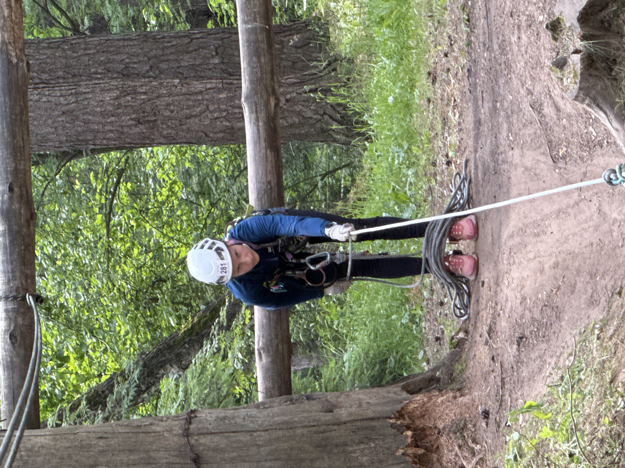 A young person wearing a white helmet with the number 281, a blue long-sleeved shirt, black pants, and pink shoes is shown from the waist down, holding a coiled rope. They are on a dirt path in a wooded area. A thick wooden beam is visible in the background, supported by two wooden posts. The environment is a forest with trees and green foliage. The image is taken in Chaikovsky, Russia.