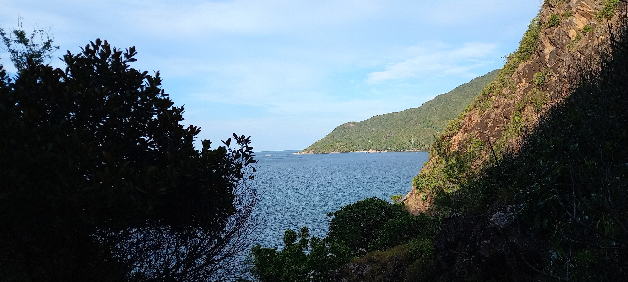 This image captures a view of the coastline near Baan Chaloklum, Thailand. The scene includes the ocean, a verdant mountainous peninsula extending into the sea, and rocky, vegetated cliffs in the foreground. Trees are visible on the left side of the image, creating a frame. The sky is pale blue with sparse clouds. The water is dark and rippled. The mountain range shows dense greenery. The rocky cliffs exhibit a mix of rock face and vegetation.
