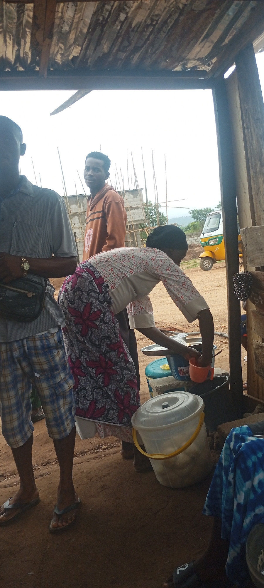 The image depicts a scene in what appears to be Zango-ndaji, Nigeria. A partially covered shelter provides framing. A woman wearing a patterned skirt and light-colored shirt leans over a bucket, using an orange cup to scoop from it. Beside her is a gray lidded bucket, a blue bucket on a scale, and other containers. Two men are standing. One is wearing a grey collared shirt and plaid shorts. The second is wearing an orange hoodie. In the background are a partially constructed building and a three-wheeled vehicle. The ground is bare earth.