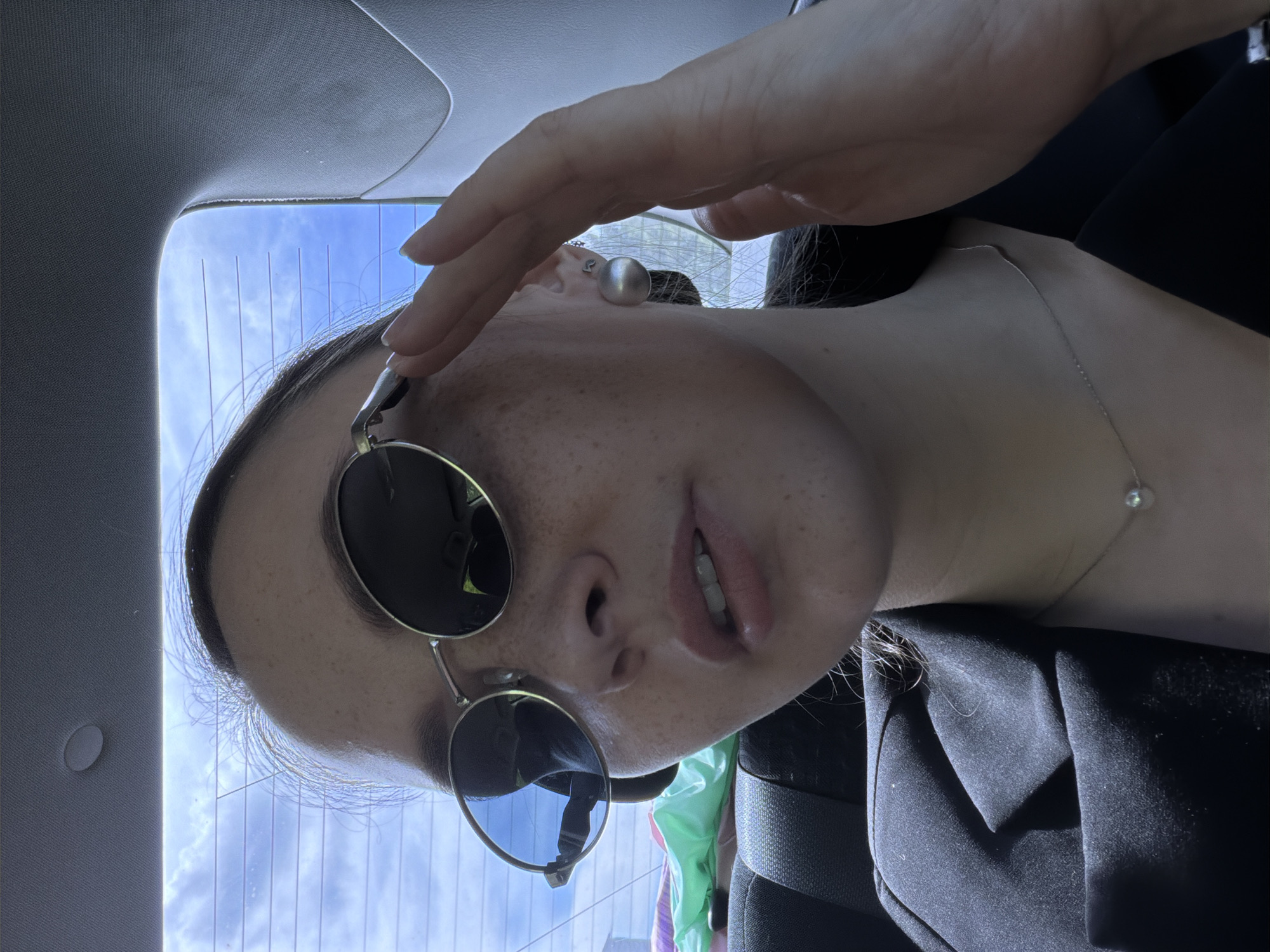 A young woman with fair skin and freckles is captured in a close-up shot, likely taken inside a vehicle, possibly on a road trip. She is wearing round, metallic sunglasses and a black blazer. A delicate silver necklace with a single pearl adorns her neck, and a large, spherical earring is visible on her left ear. Her right hand is raised, with fingers delicately touching the frame of her sunglasses. The background reveals a bright blue sky with scattered clouds, seen through the car's rear window, which has horizontal defroster lines. The interior of the car is dimly lit, with a portion of a seatbelt and some colorful items, possibly bags, visible in the backseat. There is no specific indication of the city of Chaikovsky, Russia in the image.