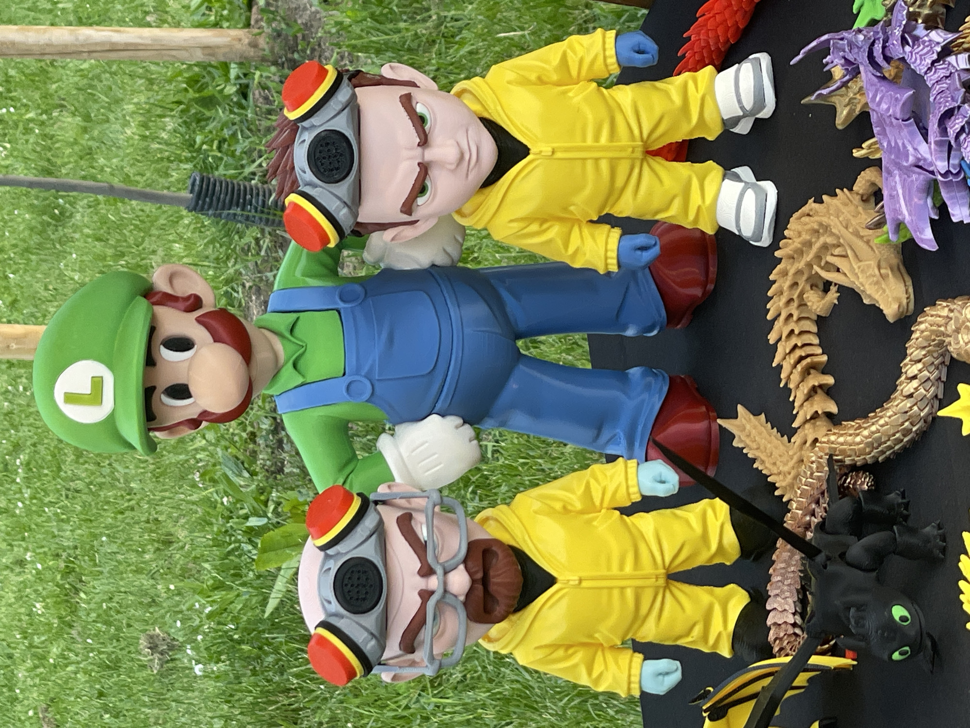 The image presents a still life arrangement of figurines positioned on what appears to be a black surface, placed outdoors on grass. The figurine set includes a depiction of Luigi from the Mario franchise, as well as two figurines resembling characters from the television series "Breaking Bad," specifically Walter White (Heisenberg) and Jesse Pinkman, characterized by their yellow hazmat suits and gas masks.
The background shows a patch of green grass, suggesting an outdoor setting. Next to the Breaking Bad characters, there is a collection of dragon figurines, varying in color from gold to purple to black.
While it's difficult to pinpoint the exact location of Zemun Urban Municipality, Serbia, from visual data alone, the grass and outdoor setting indicate that this arrangement could be displayed in a park, garden, or other outdoor public space in Zemun.