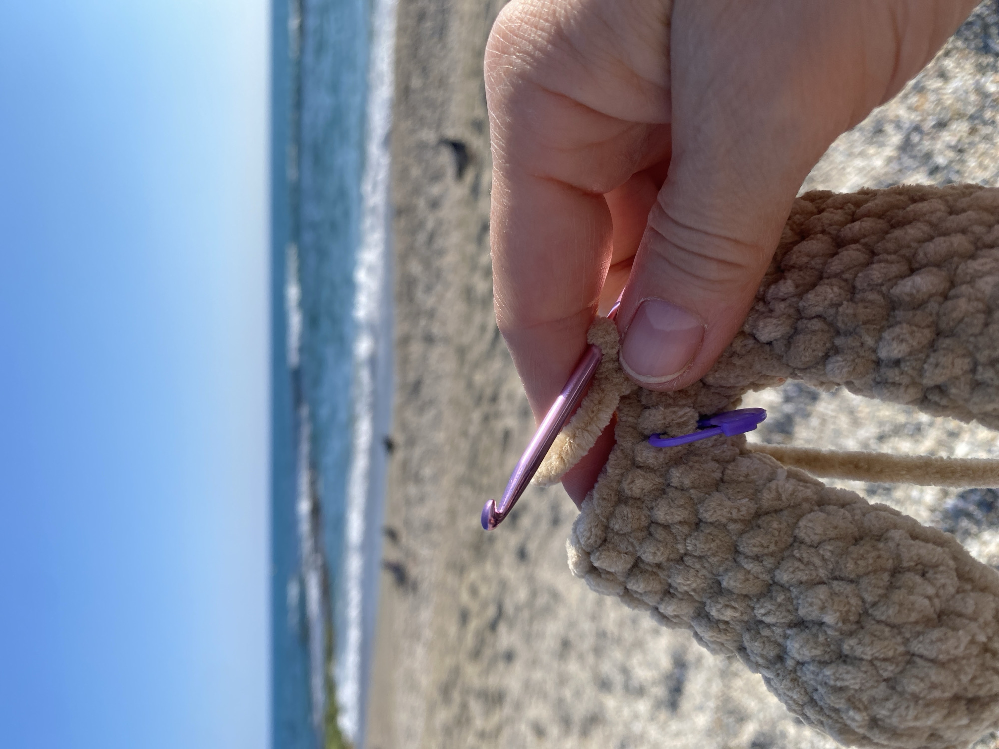 The image shows a close-up of a person's hand holding a partially crocheted, stuffed object made of beige chenille yarn. A pink crochet hook is inserted into a stitch, and a purple stitch marker is also visible. The background shows a beach with waves and a clear blue sky. Based on the environmental context, the location is likely Alanya, Turkey, as suggested in the prompt.