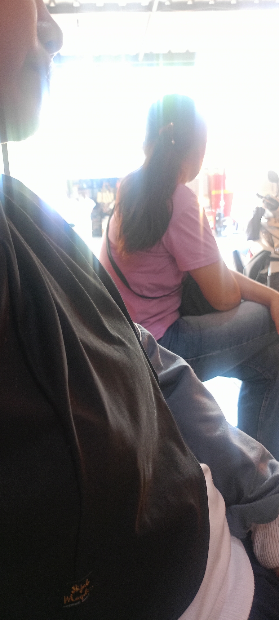 In Mekar Rahayu, Indonesia, two individuals are partially visible in a casual outdoor setting. The foreground features a person wearing a black jacket and the shoulder of their companion, who is wearing a pink t-shirt and blue jeans. The background is bright and slightly out of focus, with hints of buildings and possibly vehicles. The lighting suggests it is daytime, with strong sunlight creating glare. The overall impression is a candid snapshot of people in a relaxed, urban or semi-urban environment.