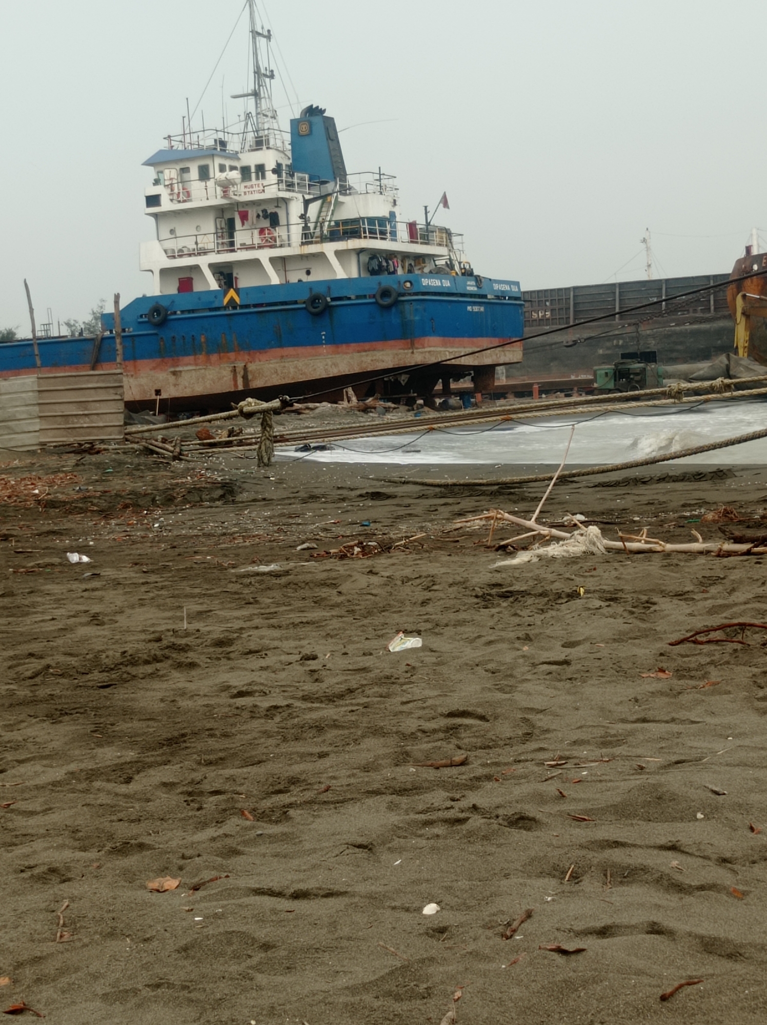 The image depicts a vessel, identified by the markings "DIPASENA DUA" and "JAKARTA INDONESIA" on its blue hull, grounded on a sandy shore. The vessel appears to be partially dismantled or undergoing repair, with exposed hull sections and visible scaffolding. In the background, other ships and industrial structures are partially visible, suggesting a shipyard or port environment. The foreground is dominated by a sandy beach strewn with debris, including wood, plastic, and natural detritus. The sky is overcast, casting a diffused light over the scene. The location is identified as Maribaya, Indonesia. There are no people visible in the image.