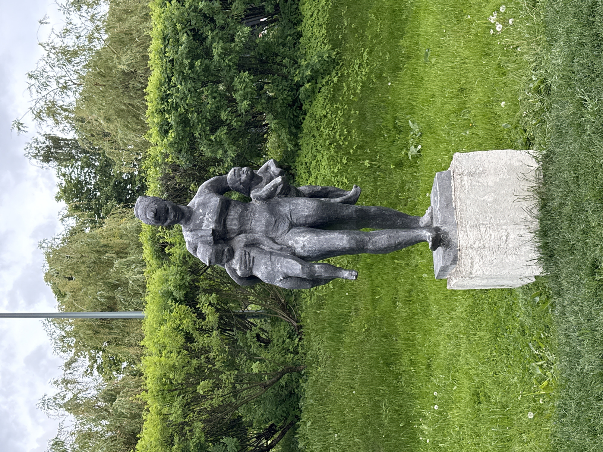 A medium, eye-level outdoor shot depicts a sculpture in a grassy park in Moscow, Russia. The sculpture is a dark, possibly bronze, human figure standing on a light grey stone plinth. The figure appears to be an adult carrying two children on its back and chest. The background is a lush green lawn with patches of white wildflowers. Behind the sculpture, there are various trees and bushes, adding depth to the scene. The sky is visible in the upper left corner, with a mix of grey and white clouds. A light pole can also be observed in the background, left of the sculpture.