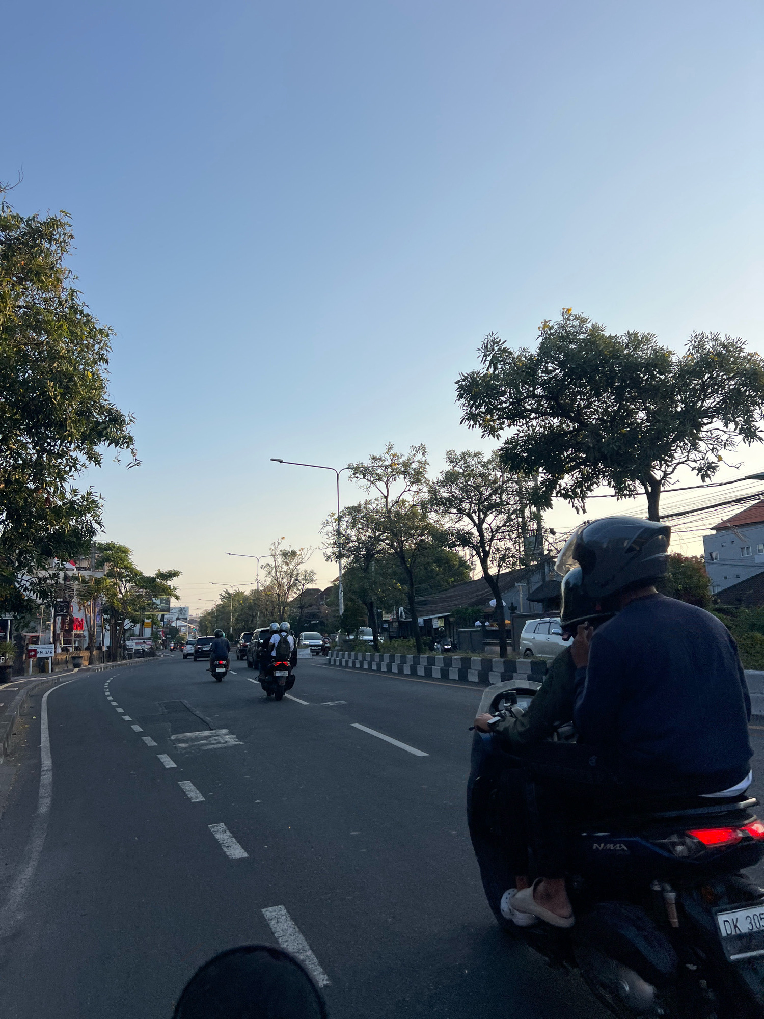 The image captures a street scene in Denpasar, Indonesia, at dusk. The perspective is from the back of a motorcycle, looking forward along a paved road with a curved lane. Several motorcycles and cars are traveling in the same direction. On the left side of the road, there are buildings, trees, and signs, including one with the word "KELUAR" (exit in Indonesian). On the right, trees and buildings are visible. The sky is clear and light blue, transitioning to a lighter hue near the horizon. The road is marked with dashed white lines. The foremost motorcycle carries two individuals wearing helmets. The license plate on the motorcycle in the foreground is partially visible and reads "DK 305".