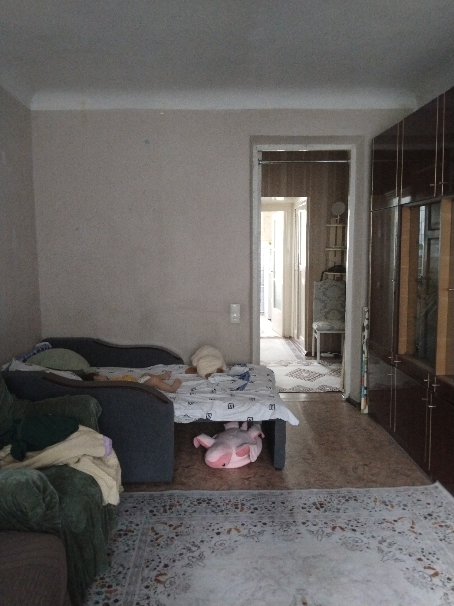 The image shows an indoor scene, likely a living room or bedroom, in a dwelling located in Ötegen batyr, Kazakhstan. In the foreground, a large, dark gray sofa is partially visible on the left, with a green armchair in front of it draped with blankets. A bed with white bedding featuring a black geometric pattern is positioned in the center of the room. On the bed, a person is lying down, wearing a yellow top and what appears to be a diaper. A large, light-colored stuffed animal is on the right side of the bed. Another pink stuffed animal is lying on the floor underneath the bed. To the right of the bed, a doorway opens into another room, where a chair with a patterned upholstery is visible. Further to the right is a tall, dark wood cabinet with multiple doors and drawers. The flooring consists of a light brown patterned linoleum and a cream-colored rug with a floral pattern in the foreground. The walls are painted a neutral color, and the ceiling is visible at the top of the image. Natural light enters the room from the doorway in the background, illuminating the scene.