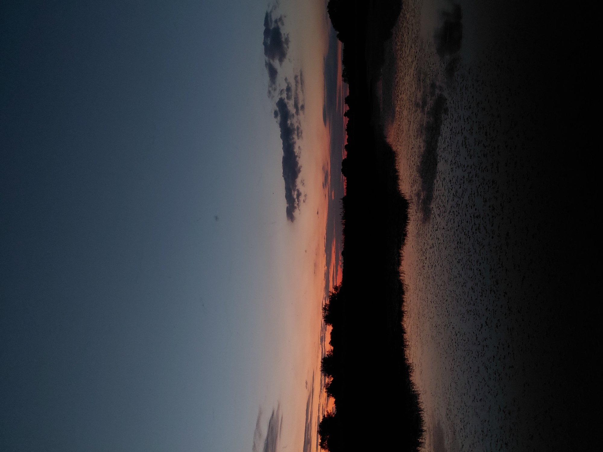 The image shows a serene sunset over a body of water, likely a lake or river, with a silhouetted tree line in the distance. The sky transitions from a deep blue at the top to shades of orange and pink near the horizon, indicative of twilight. Wisps of clouds are scattered across the sky, some catching the fading sunlight. The water's surface is dark and reflects the colors of the sky, with small ripples disrupting the reflection. The overall scene is peaceful and natural, suggesting a rural or semi-rural setting. This image could be located near the village of Ustyvytsia, Ukraine, a place known for its natural landscapes.