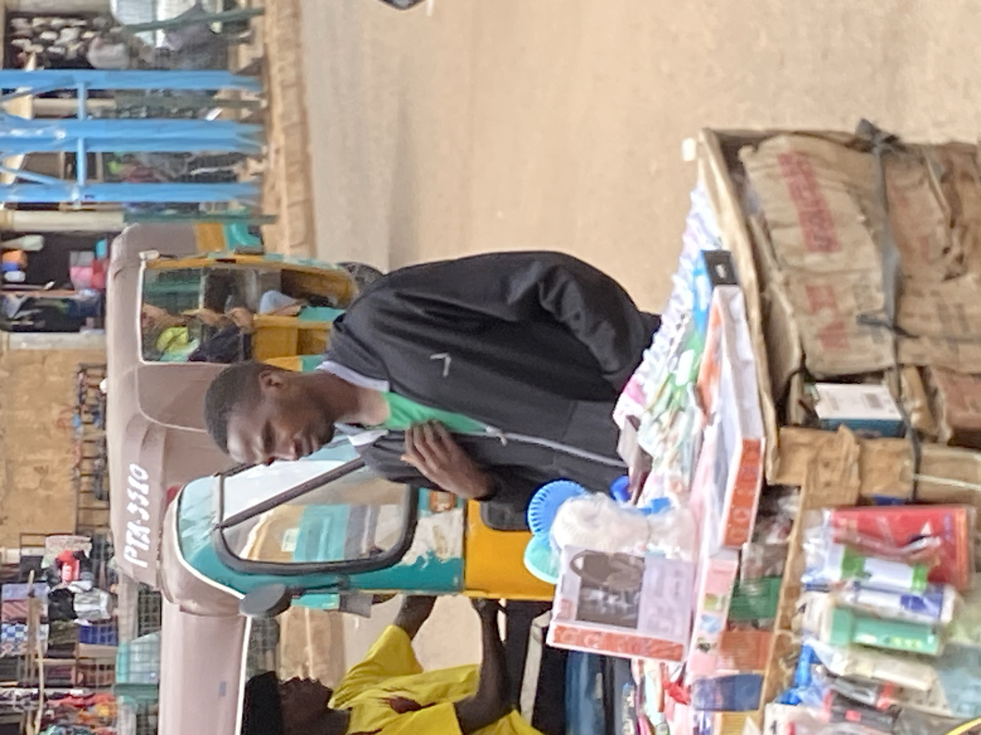 The image captures a vibrant street market scene, likely in Bukuru, Nigeria, under clear daylight. In the foreground, a man with short dark hair, wearing a black hooded jacket over a green shirt, is attentively looking down at a vendor's stall to his right. His posture suggests he is browsing or interacting with the goods.
To the man's left, a cream and teal-colored auto-rickshaw, commonly referred to as a 'keke napep', is a prominent feature. The vehicle has "PTA-3310" clearly visible on its side. Partially visible beneath the auto-rickshaw and near the stall is another person wearing a bright yellow top. A third individual can be seen inside the auto-rickshaw.
The vendor's stall is richly stocked with various items, including numerous packaged goods that appear to be small consumer products or snacks. Behind these, large sacks with indistinct red text are stacked. The background reveals more of the bustling market environment, with blue metal shelves or racks and other market structures, hinting at a lively commercial area. The overall scene depicts a typical daily market activity with no strong emotions discernible from the visible individuals.