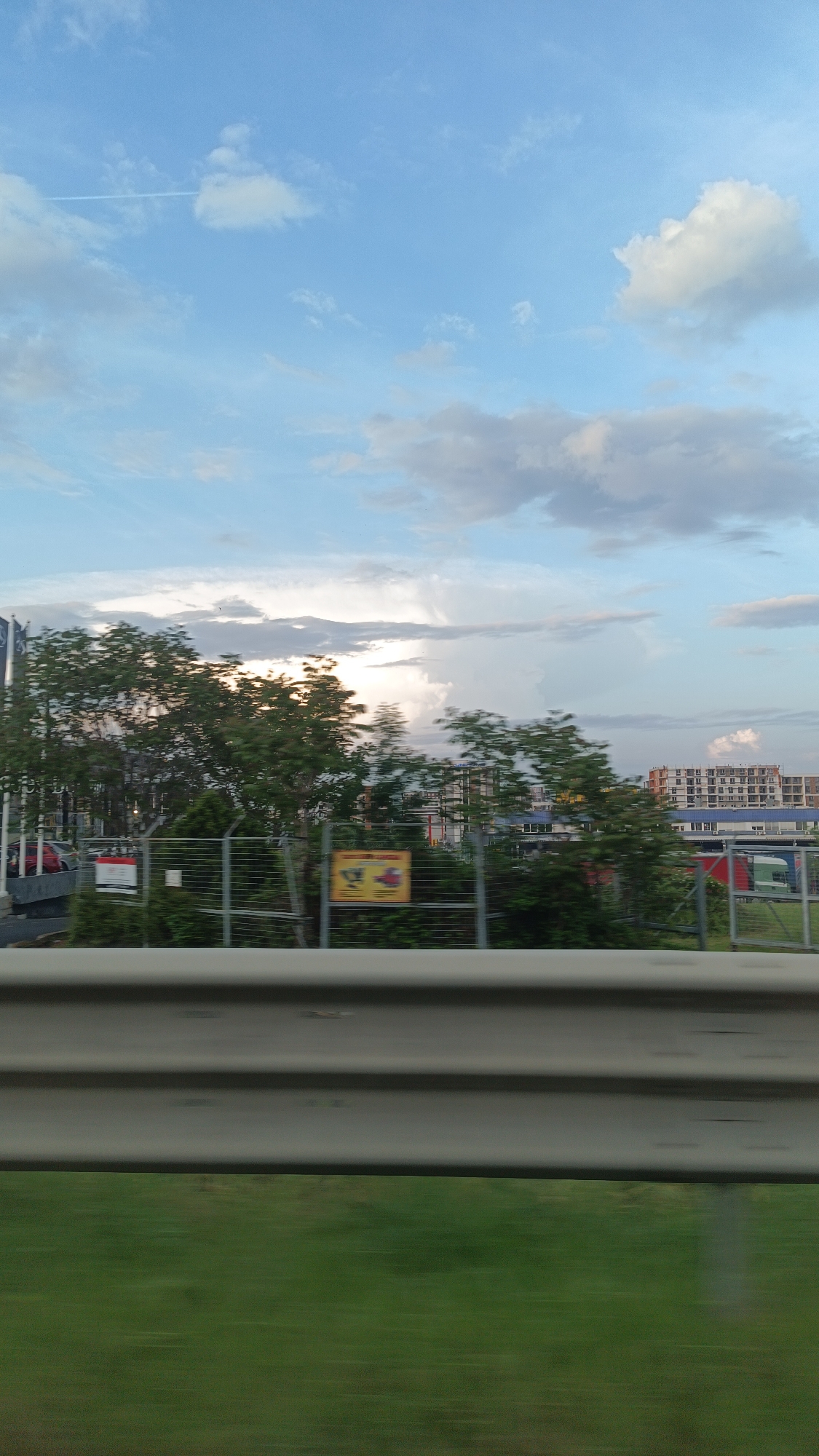 The image shows a view from a moving vehicle. A grey metal guardrail is in the foreground. Beyond the guardrail is green vegetation, likely a grassy area. Behind the vegetation, there is a wire fence. Behind the fence are trees and, further back, buildings, including what appears to be a multi-story residential building with a brick facade. A sign with yellow and red colors is partially visible on the fence. The sky is visible above the trees and buildings, featuring blue sky and white clouds, including a contrail. Considering the type of construction in the background and the overall feel of the landscape, it is plausible the location is near a highway or main road on the outskirts of Burgas, Bulgaria, but there is nothing to definitively confirm that. No people are visible in the image. The scene is taken during daytime.