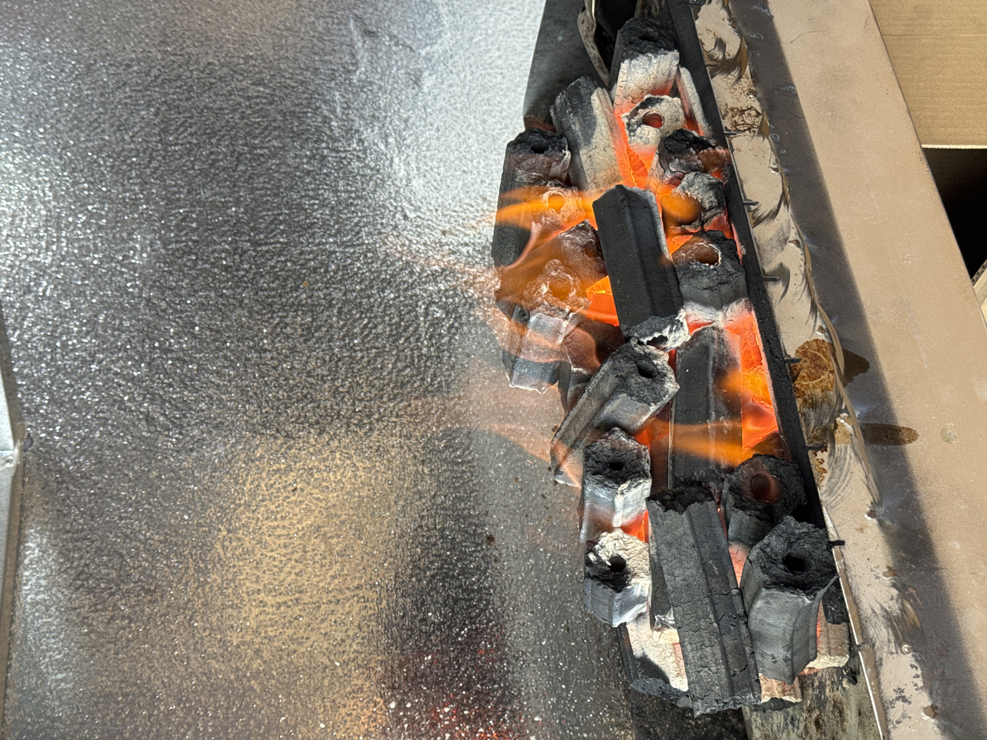 The image shows a close-up view of charcoal briquettes burning in a grill. The briquettes are hexagonal in shape and are stacked on top of each other, with flames and glowing embers visible between them. The background is a textured, metallic surface that appears to be part of the grill's interior or a surrounding structure. The lighting is warm, highlighting the intensity of the fire. The scene is devoid of any people or specific landmarks that would indicate a location within Nha Trang, Vietnam. It is a general depiction of a barbecue setup.