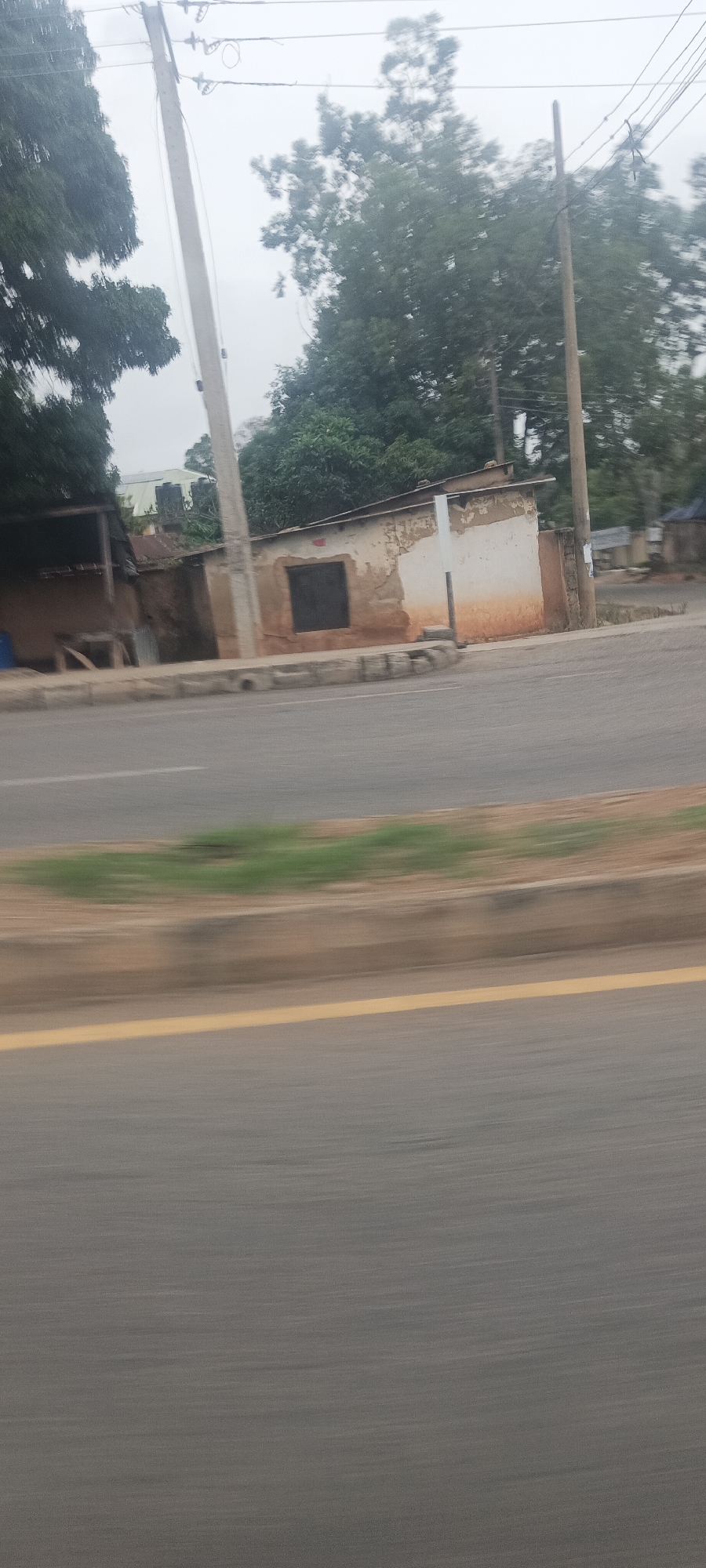 The image captures a motion-blurred roadside scene in Jos, Nigeria, suggesting it was taken from a moving vehicle. In the foreground, a gray asphalt road with a distinct yellow median line occupies the lower half of the frame.
Above the road, a concrete curb separates it from a narrow dirt and grass verge, also exhibiting motion blur. Several tall utility poles, characteristic of power or communication infrastructure, stand along the roadside, carrying a network of wires.
In the mid-ground, a cluster of simple, utilitarian structures is visible. On the far left, a dark, open-sided stall or shelter with a corrugated metal roof is partially seen. Adjacent to it is a small, single-story building with textured, earthy-toned walls showing signs of wear and peeling plaster, revealing a darker underlayer. A dark, rectangular opening, likely a closed window or door, is centrally placed on its facade. Further to the right, another similar, weathered structure is partially visible. The roofs of these buildings appear to be made of corrugated metal.
The background is filled with lush, green foliage from numerous trees, some appearing blurred. The sky above is uniformly overcast, indicating a cloudy day. There are no visible people, discernible activities, or any text within the frame. The scene portrays an everyday street view in a semi-urban or developing area.