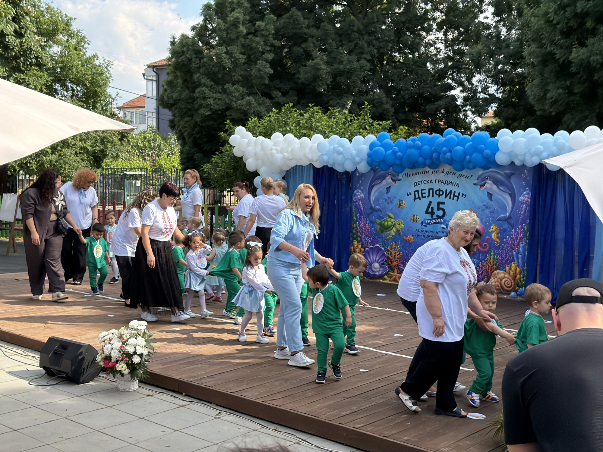 This vibrant image depicts an outdoor children's event, likely a celebration or performance, held on a wooden stage in Burgas, Bulgaria. The setting is bright and sunny, indicating daytime.
Numerous people are visible, including many young children and several adults. The children, mostly toddlers or preschoolers, are dressed in coordinated outfits: many wear bright green tracksuits adorned with a seahorse emblem, while some girls wear light blue dresses. They are arranged in lines on the stage, guided by adult women who are dressed predominantly in white tops, some with scarves, or light blue attire. One older woman in a white embroidered top and black pants is assisting children on the right side of the stage. A man in a black cap is partially visible in the right foreground, observing the event.
The children appear to be engaged in a performance or parade, holding hands and moving purposefully across the stage. The adults seem to be teachers or caregivers, attentive and supportive.
The stage features a colorful blue backdrop decorated with an underwater theme, including dolphins, jellyfish, sea turtles, and corals. Above the backdrop, an impressive arch of white and blue balloons creates a festive atmosphere. To the left of the stage, a large white awning or tent provides shade. In the background, lush green trees are abundant, with some buildings visible behind them. A large black speaker and a floral arrangement with white and pink flowers are placed on the ground next to the stage.
Prominent text on the blue backdrop, written in Cyrillic, reads: "Честит рожден ден!" (Happy Birthday!), "ДЕТСКА ГРАДИНА" (Kindergarten), "ДЕЛФИН" (DELFIN - Dolphin), and "45 години" (45 years). This clearly indicates the event is the 45th anniversary celebration of the "Delfin" Kindergarten. The overall mood is joyful and celebratory.