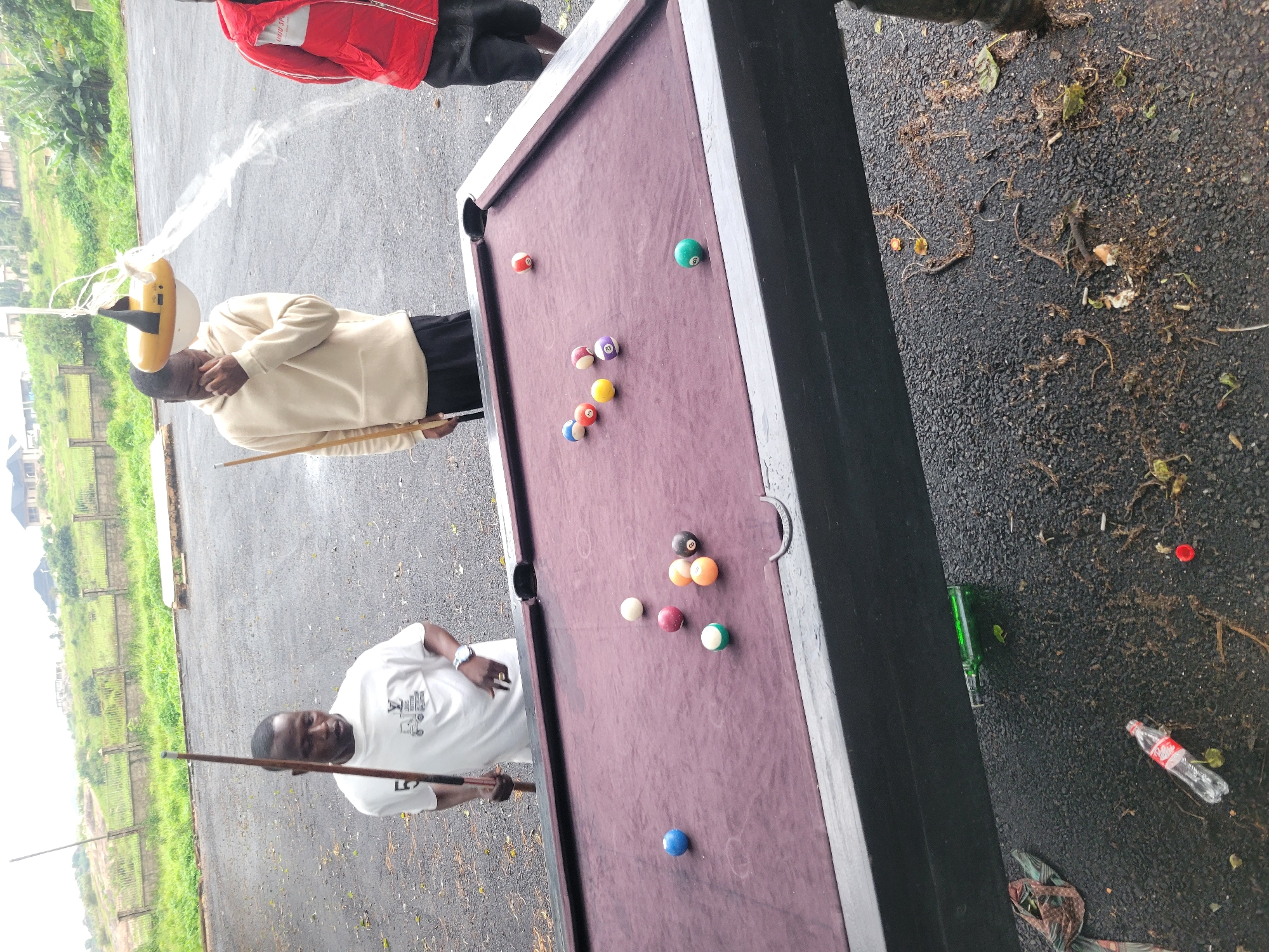 This image depicts an outdoor scene, possibly in Jos, Nigeria, featuring a group of individuals near a pool table. Three men are visible. The man on the left, wearing a white t-shirt, holds a pool cue. In the center, a man in a beige hoodie covers his face with his hand. To the right, a man in a red jacket stands. A pool table with several balls is set up on a dark asphalt surface, which is littered with debris including leaves, dirt, and what appears to be a plastic bottle and a green glass bottle. The background shows a fence, grassy slopes, and buildings in the distance, suggesting a semi-urban or suburban environment. The overall atmosphere is casual, with the pool table being the central object of interest.
